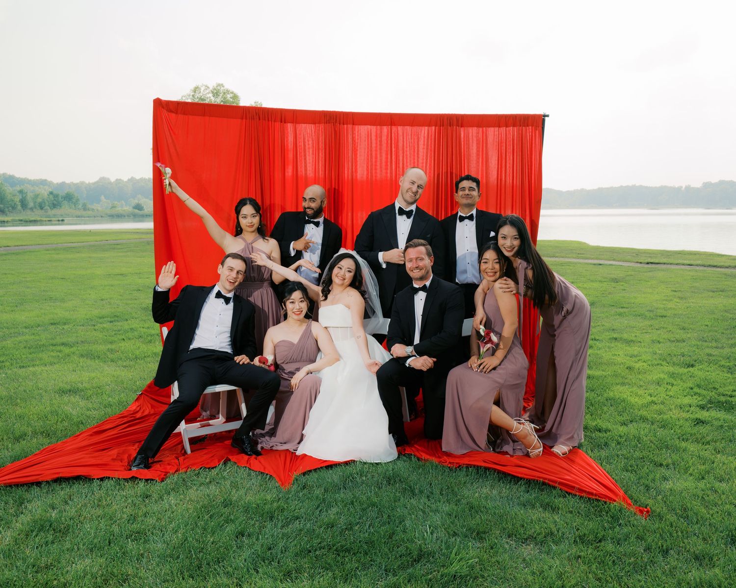Wedding party poses together against a dramatic red backdrop outdoors by a lake.