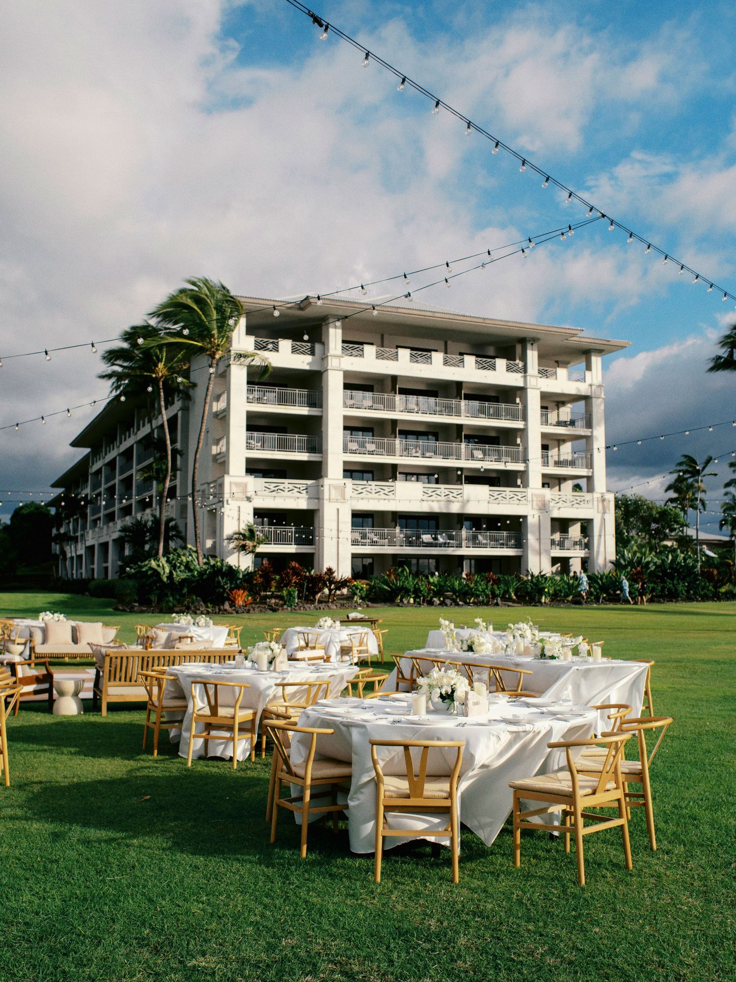 Outdoor wedding reception setup with round tables and chairs on a lawn in front of a modern beachfront resort hotel.