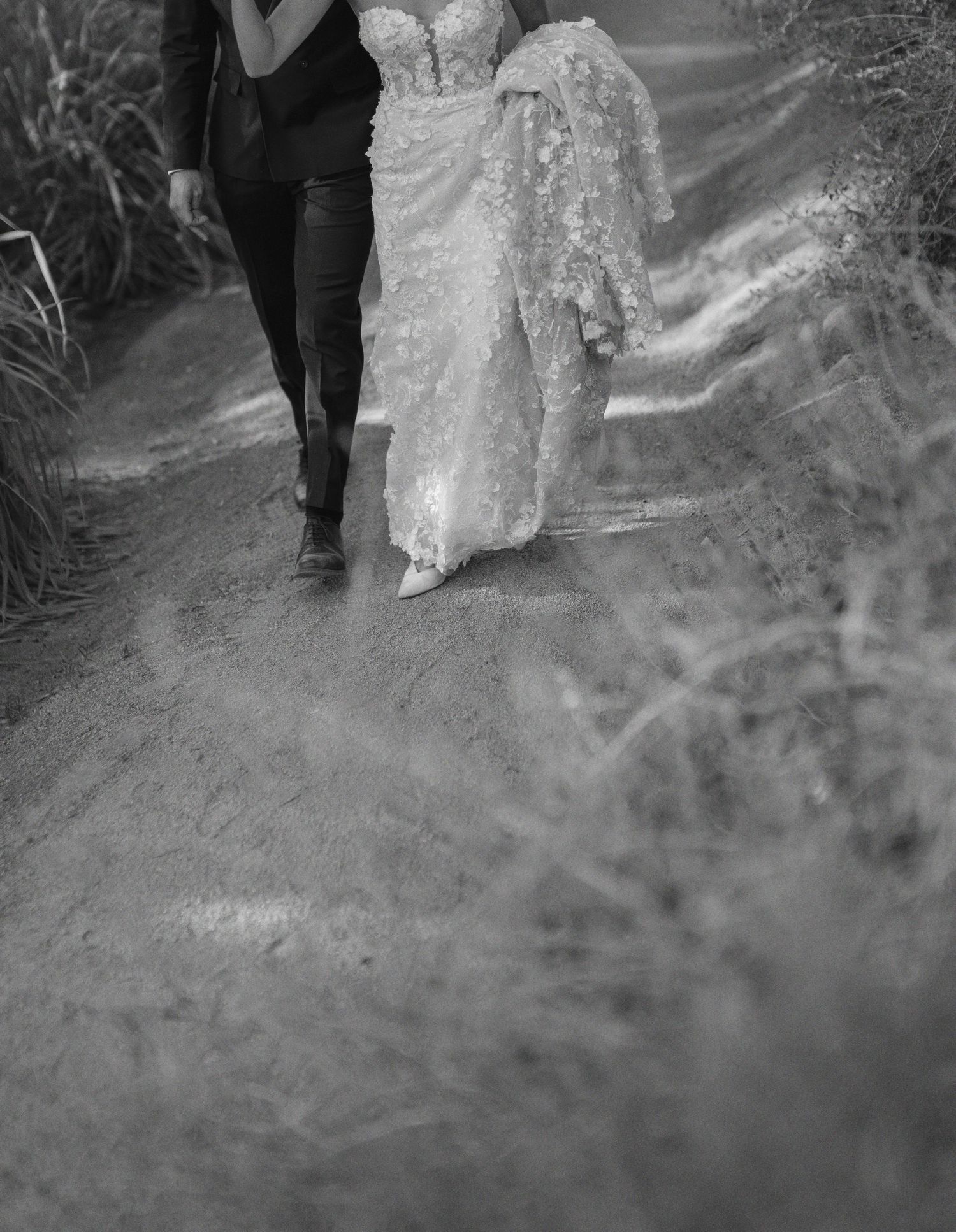 A black and white wedding photo showing a couple walking down a dirt path with a lace dress trailing behind.