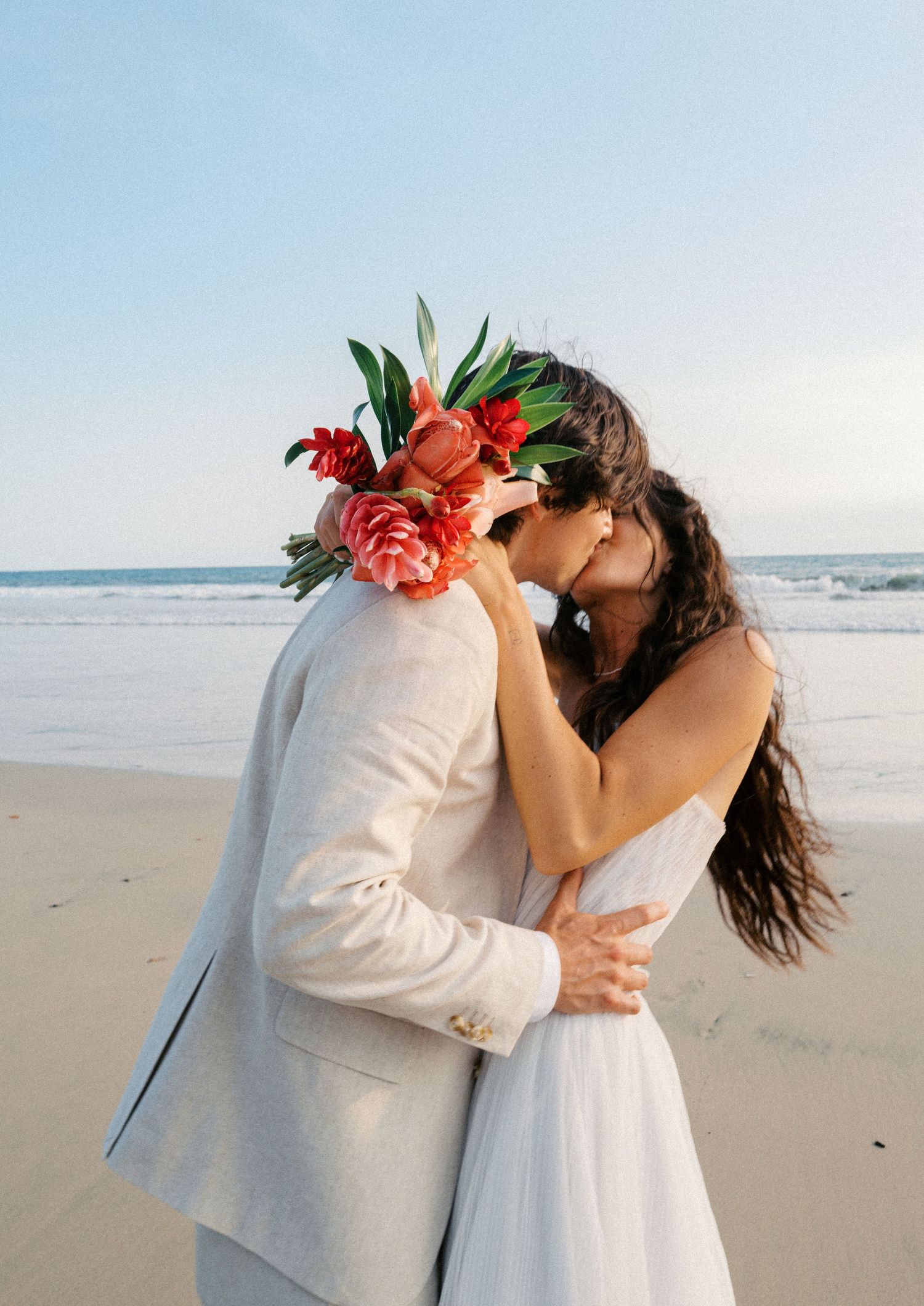 A couple in white attire embraces on a serene beach with coral-colored tropical flowers in their hair.