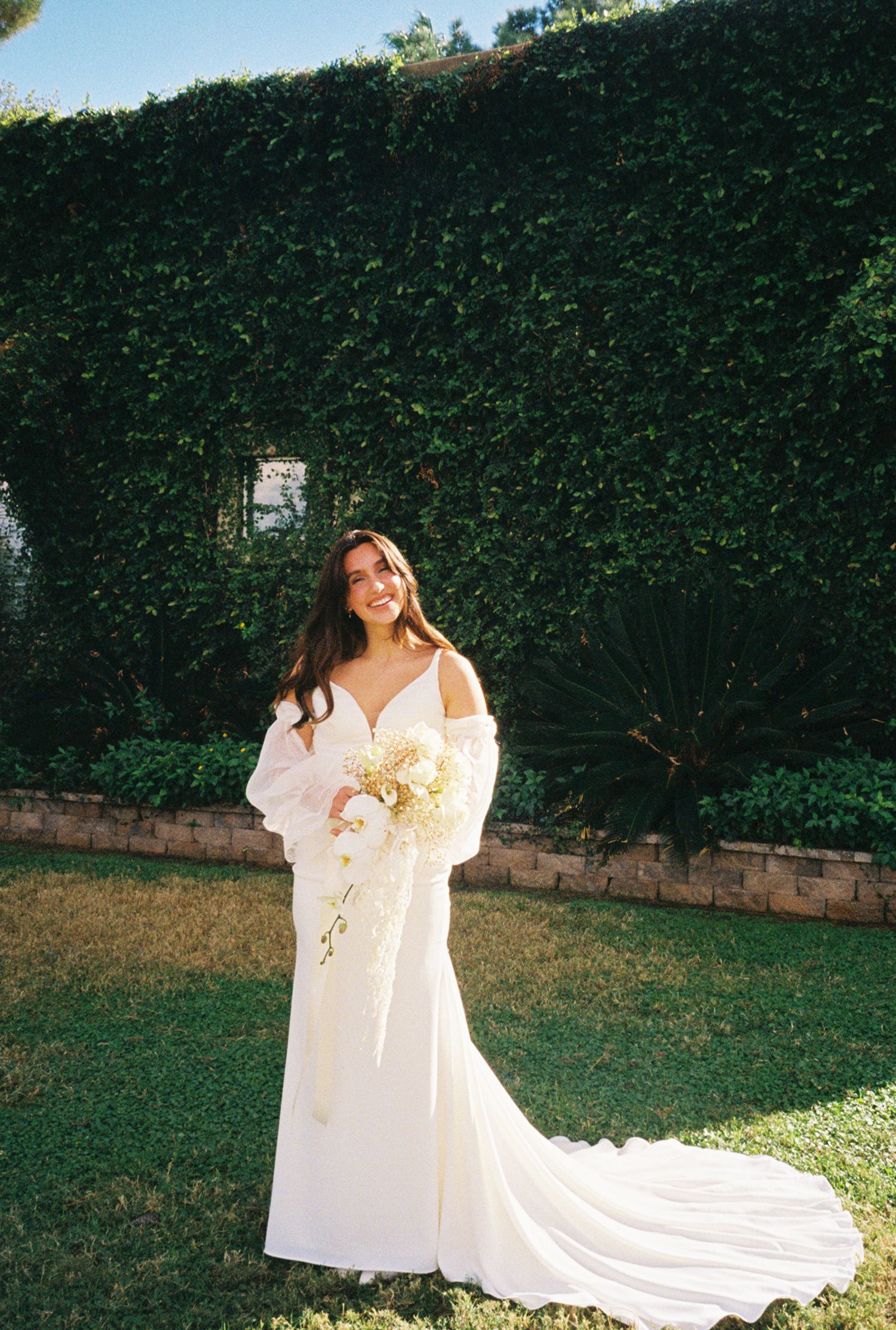 A flowing white wedding dress with long train laid out on grass against a green hedge backdrop in outdoor setting.