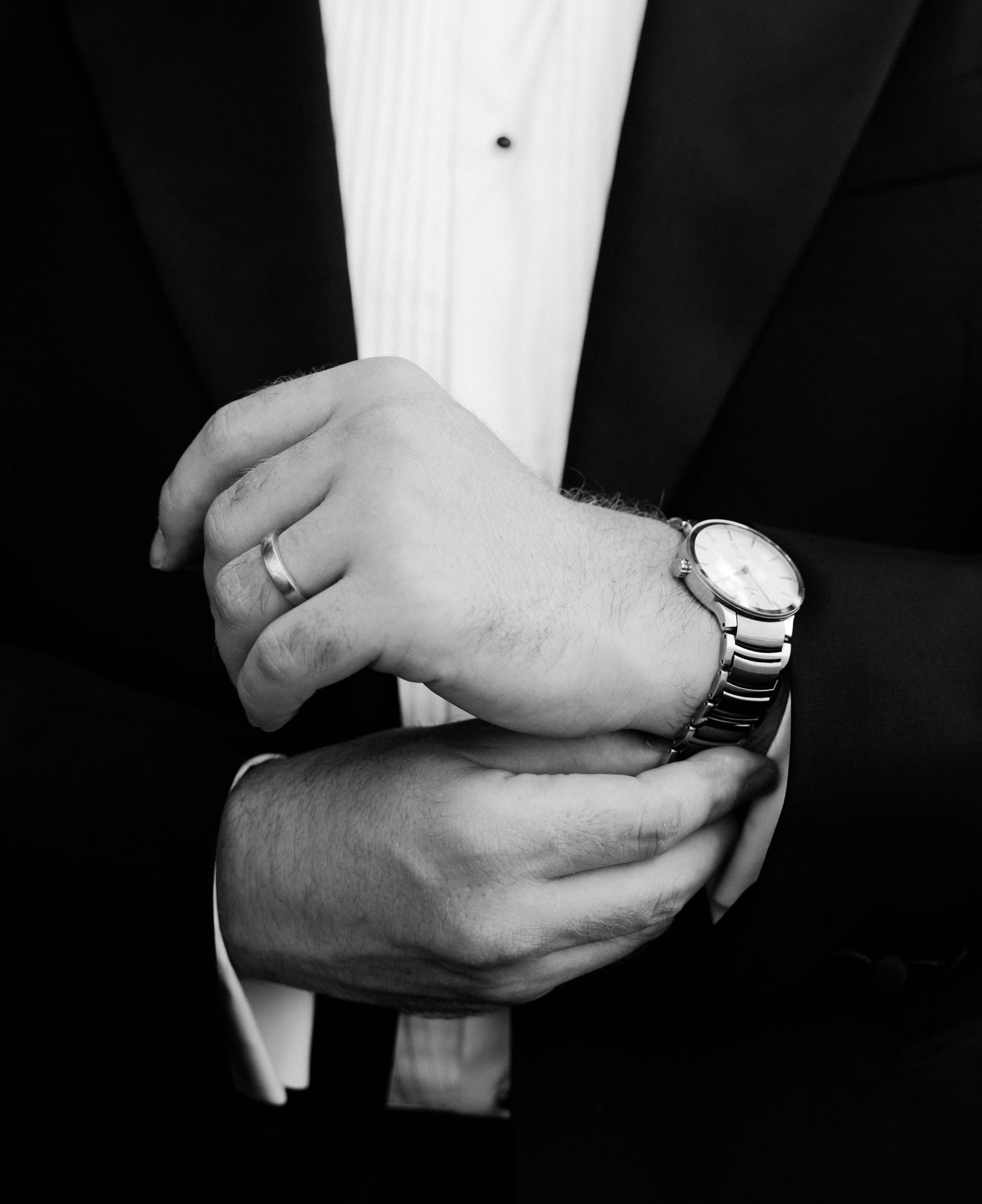 Close-up black and white photo of hands adjusting a watch while wearing a formal tuxedo and wedding ring.