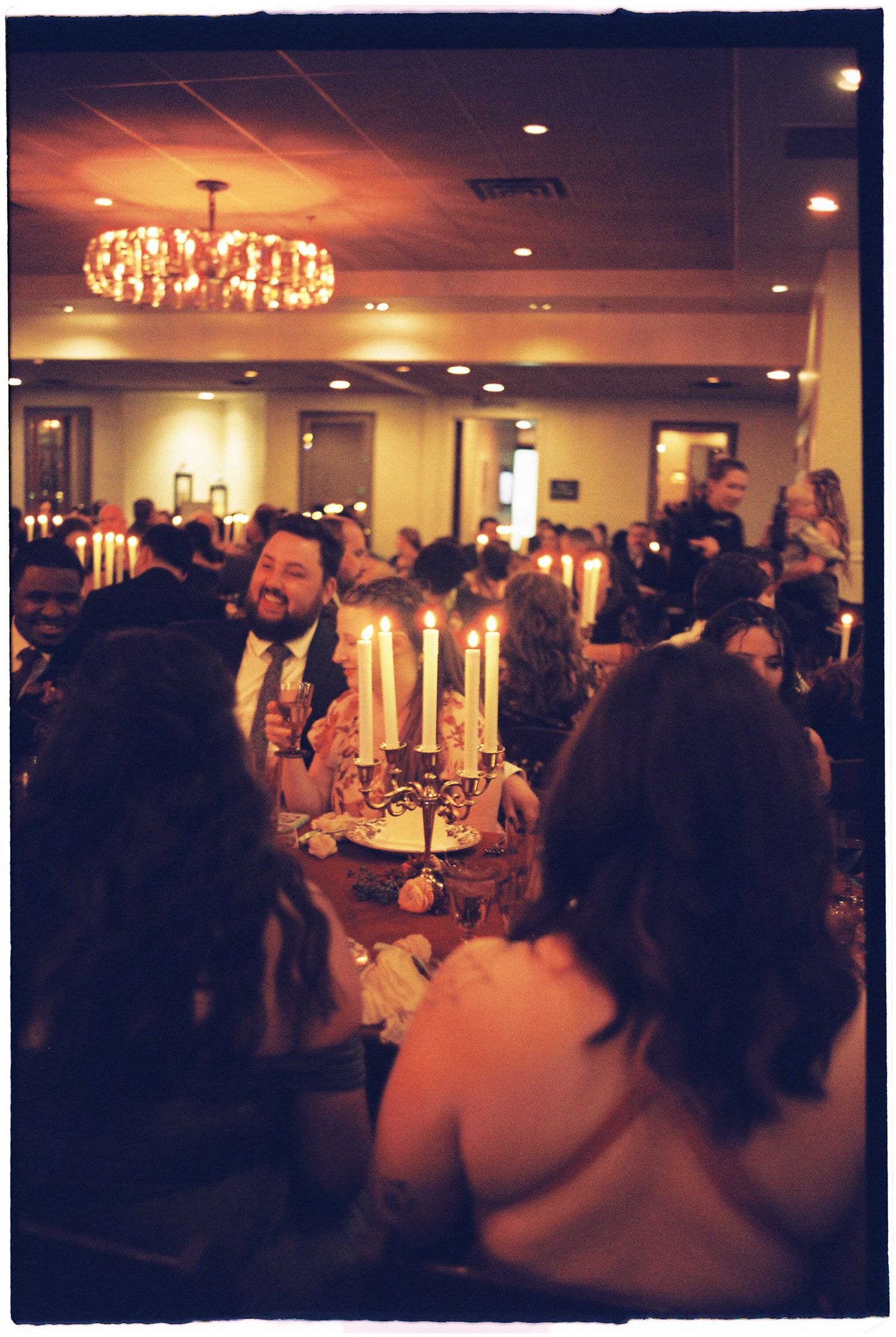A dimly lit reception hall with elegant chandeliers and guests mingling at tables during an evening celebration.