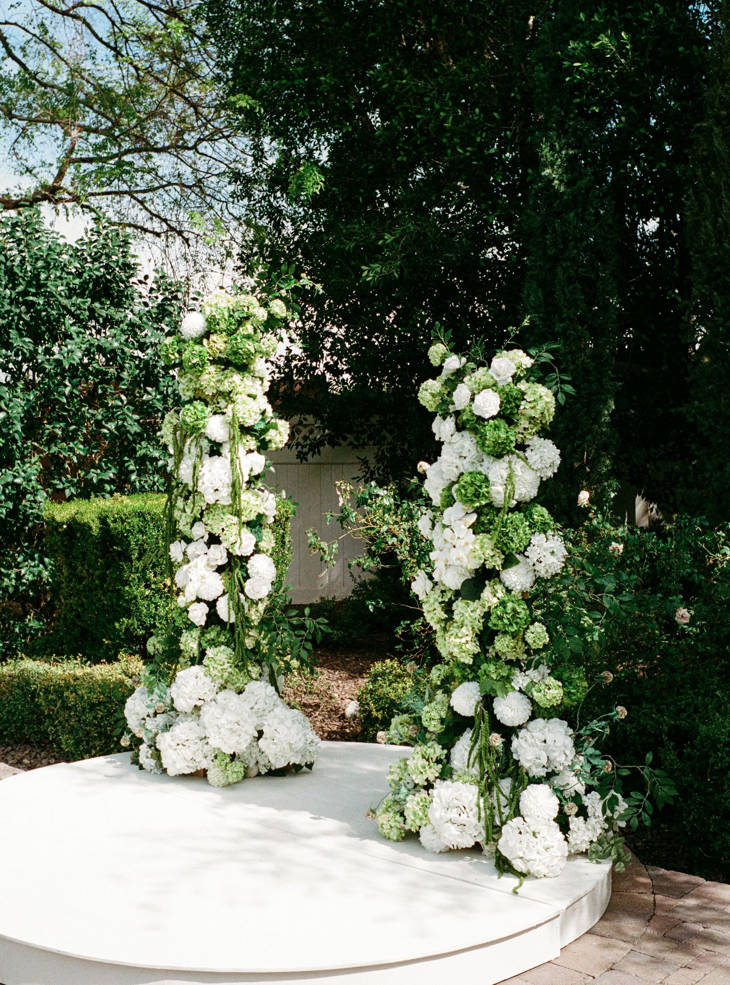 Elegant white floral arch with hydrangeas and roses decorates an outdoor garden ceremony space on a circular platform.