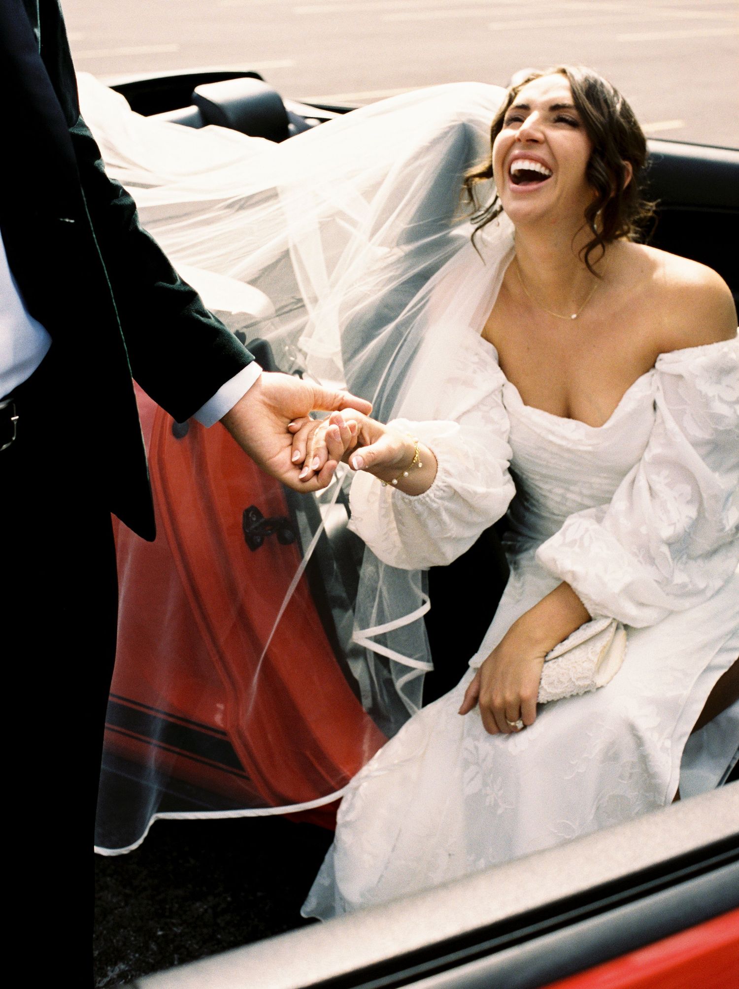 Happy newlyweds laughing and celebrating in a convertible car with white wedding dress flowing.