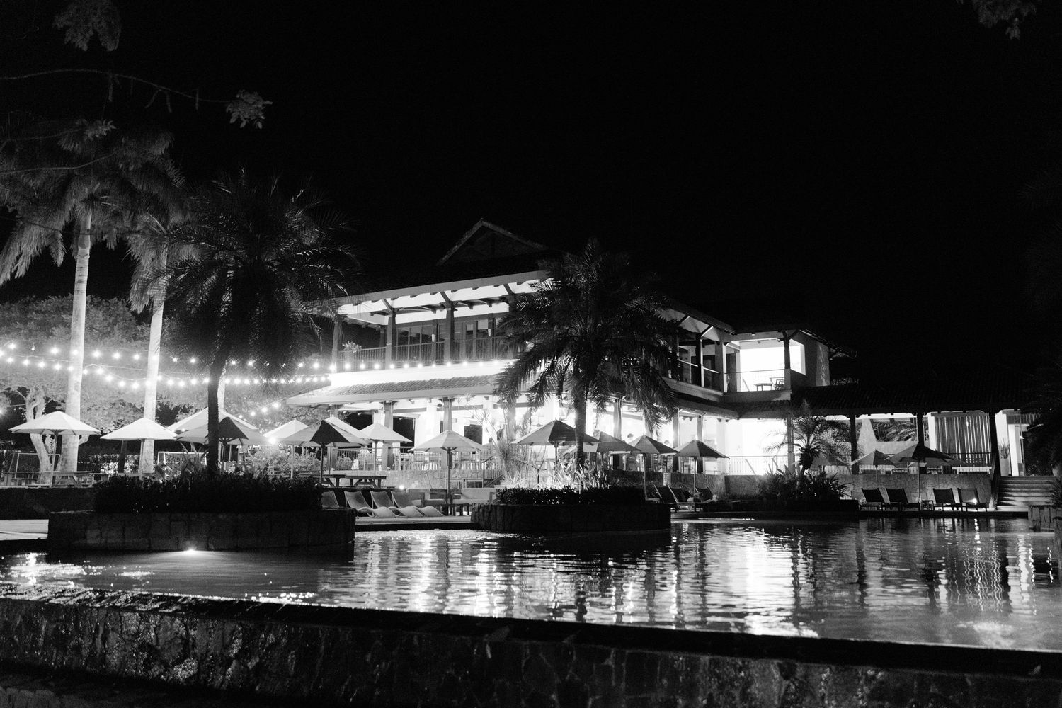 A black and white nighttime photograph of a waterfront restaurant with palm trees and reflections on the water.
