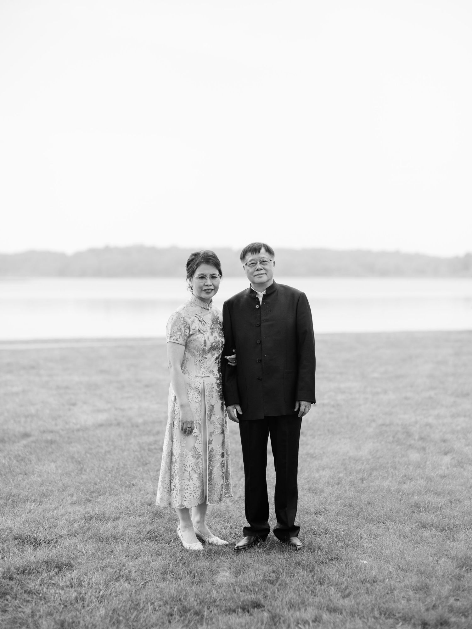 A vintage black and white photograph of a couple standing in a grassy field by the water.