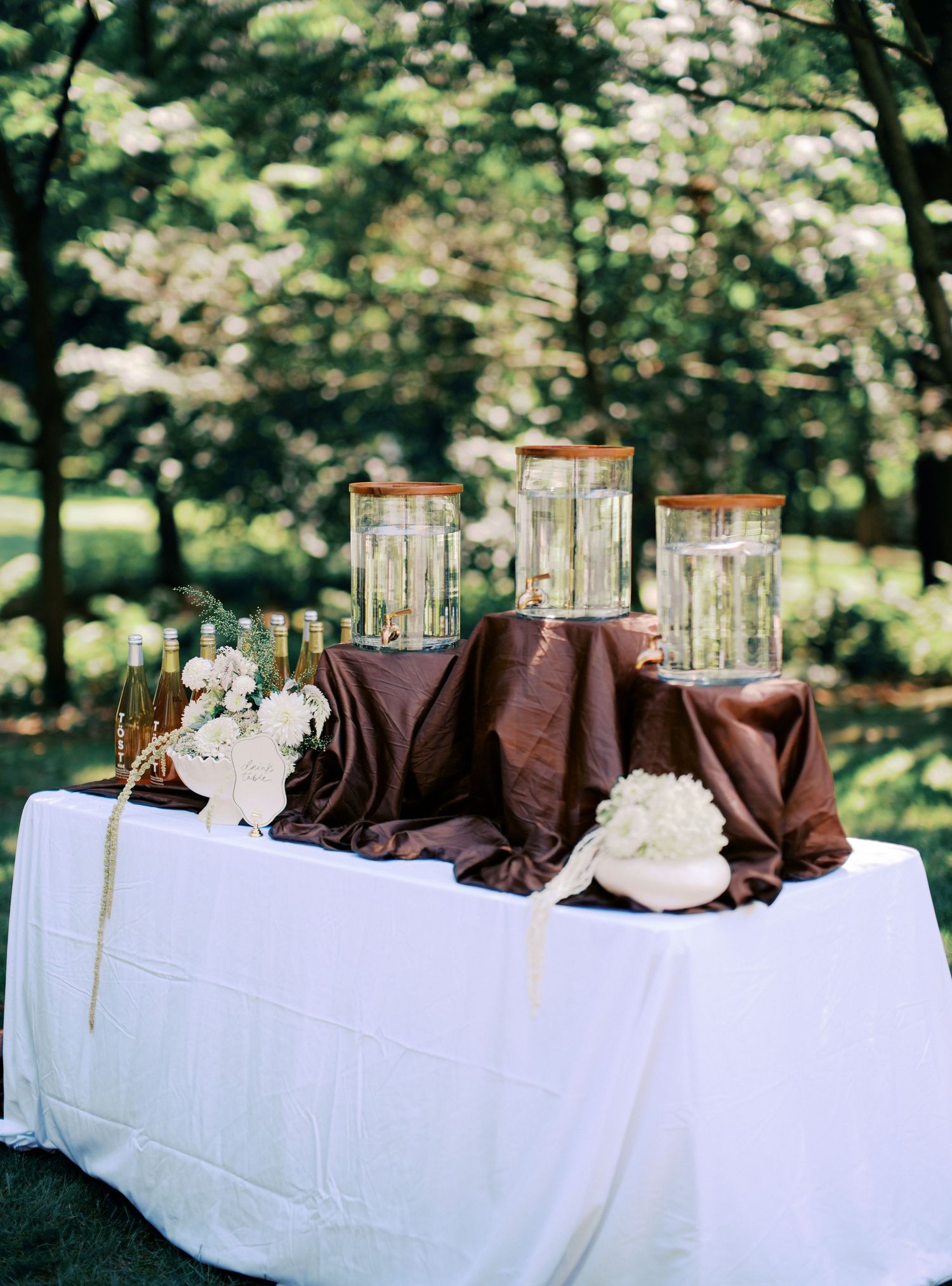 Wedding reception table set with white linens and glass candle holders in an outdoor setting.