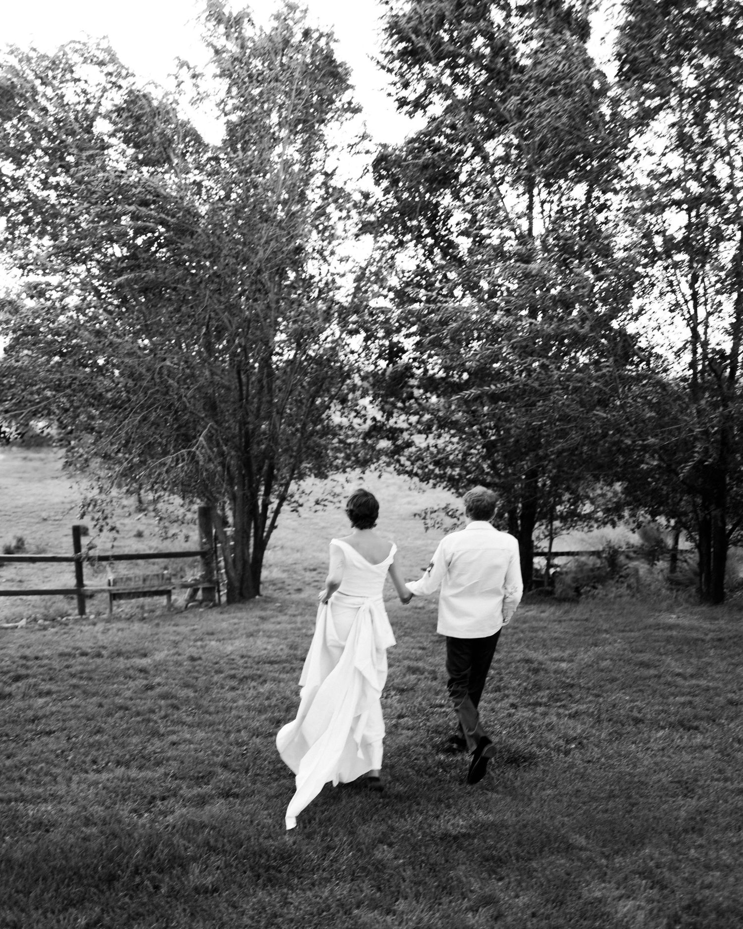 A couple walks hand in hand through a scenic meadow with trees in black and white photography.