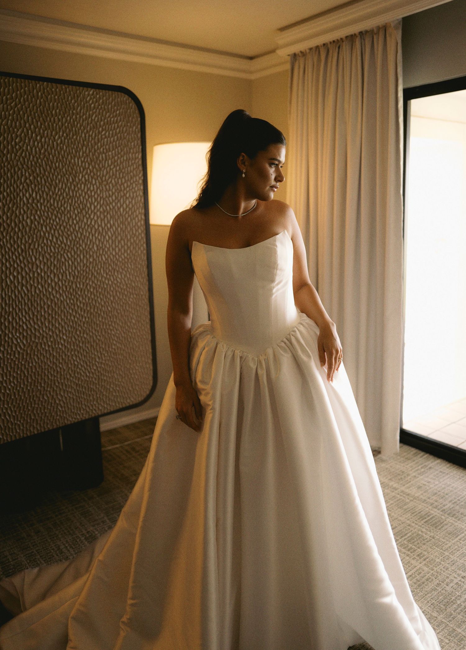 A bride in a flowing white wedding gown is photographed in a sunlit hotel room from different angles.