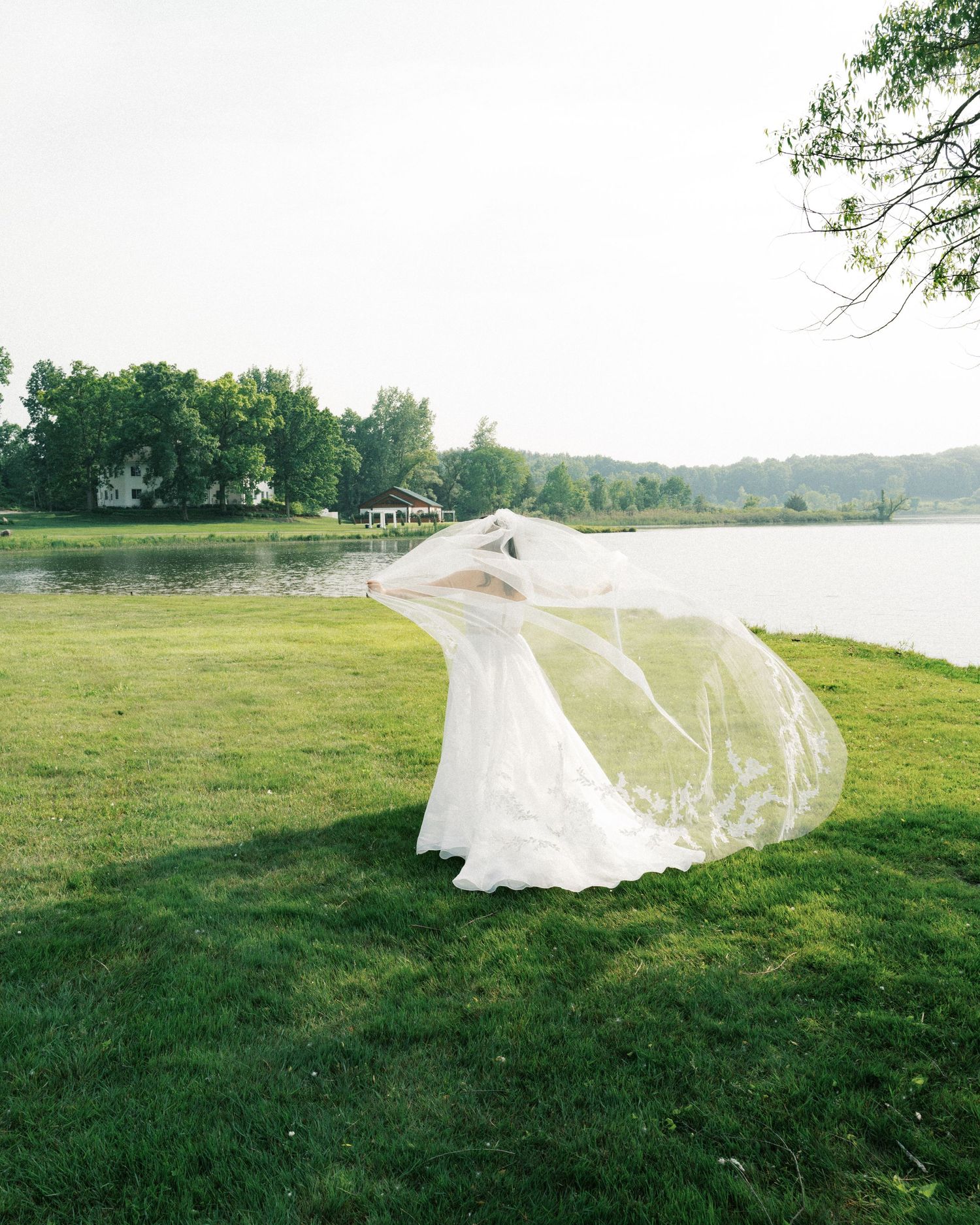 A flowing white wedding veil catches the breeze on a grassy lawn beside a lake.