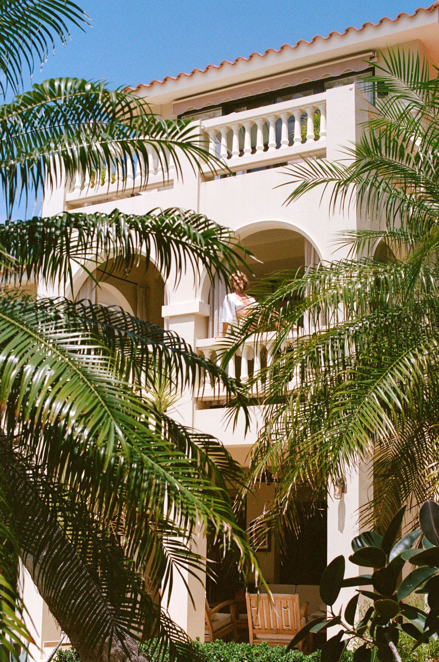 Palm trees frame the white stucco arches and balconies of a Mediterranean-style building in warm sunlight.