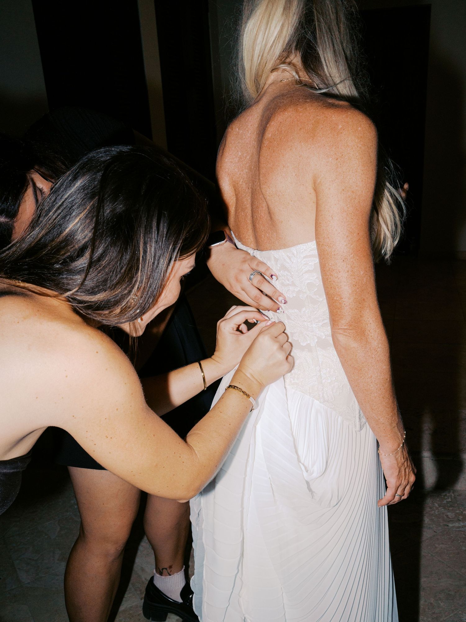 Woman in white dress being helped with zipper closure by another person in dimly lit setting.