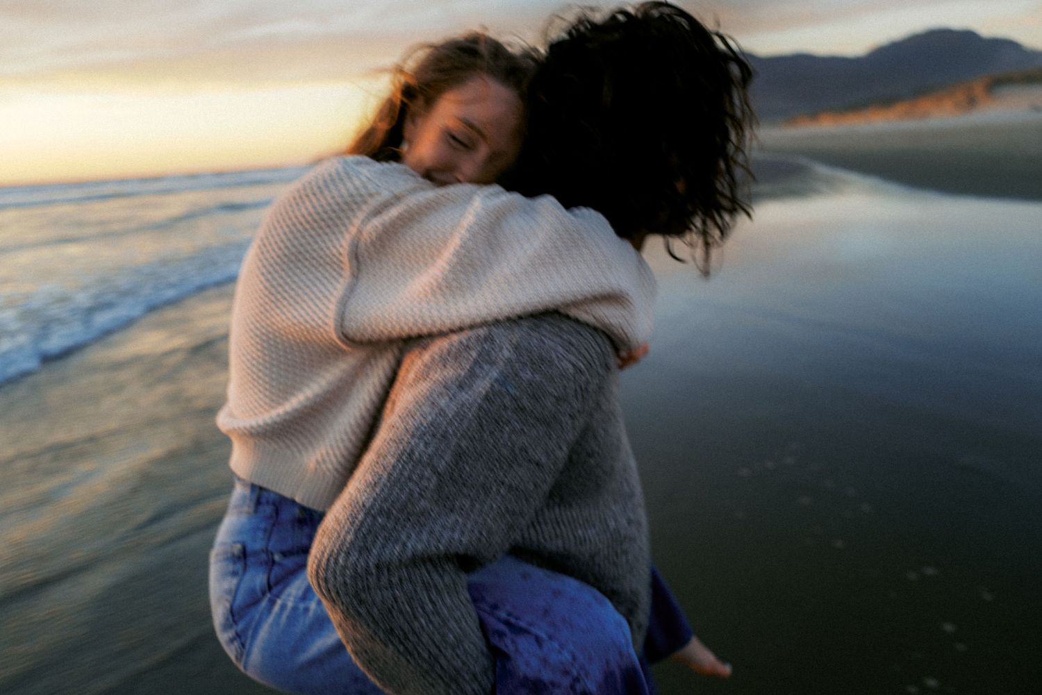 Two women share a warm embrace on a scenic beach at sunset, wearing cozy knit sweaters.