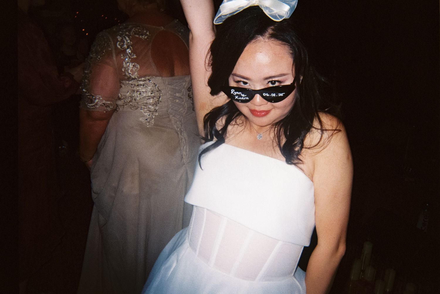 A figure in a white dress and sunglasses poses in a dark room with a flash photograph effect.