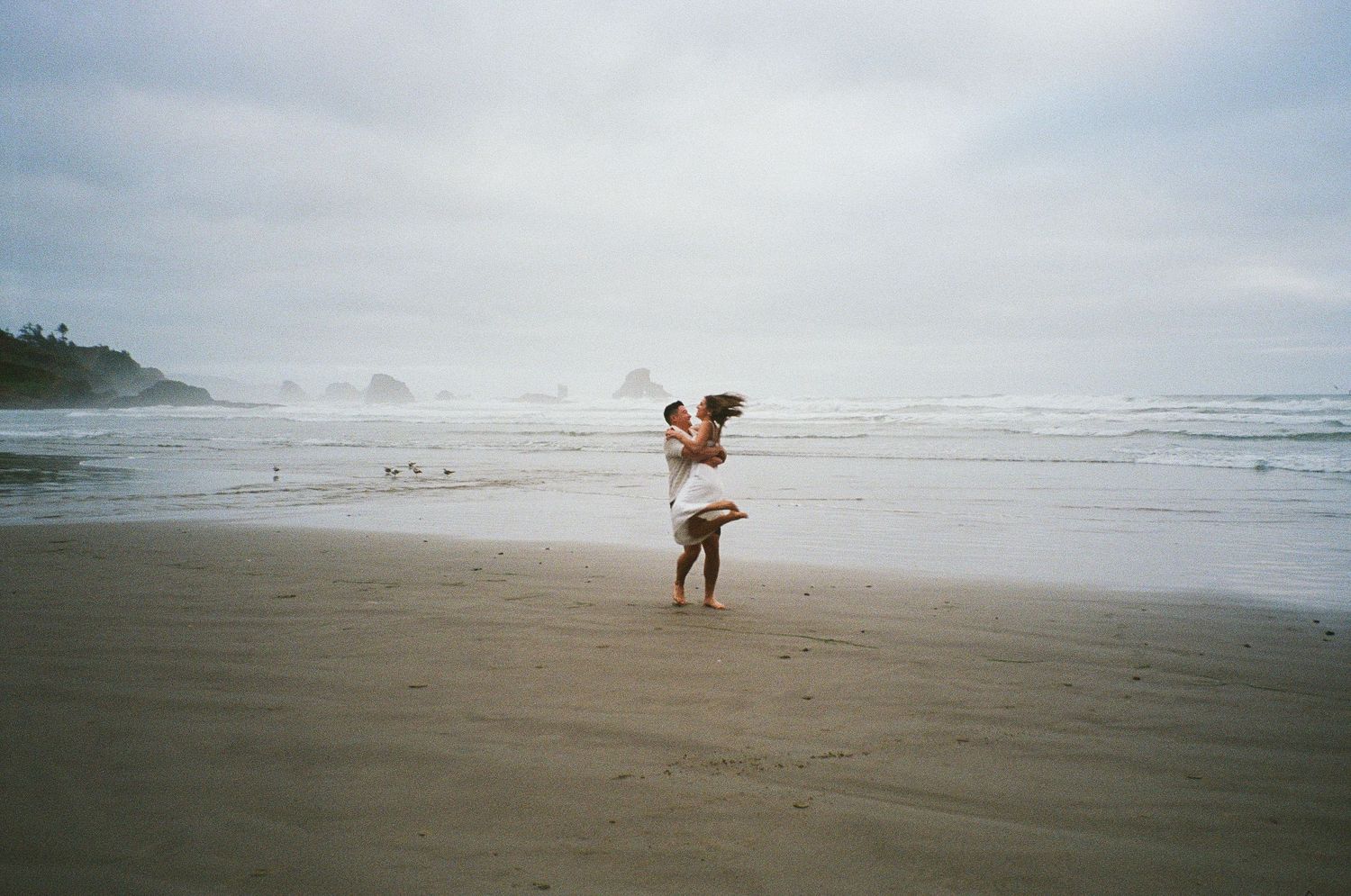 A lone figure stands on a misty beach carrying a small dog while waves roll in along the shoreline.