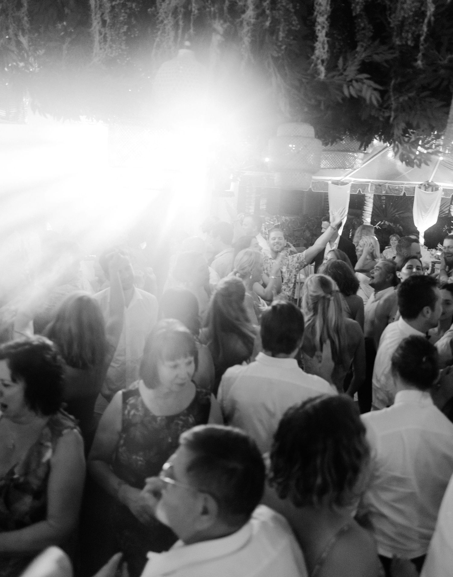 A black and white photo shows a crowded dance floor with people dancing under bright lights at a party or celebration.