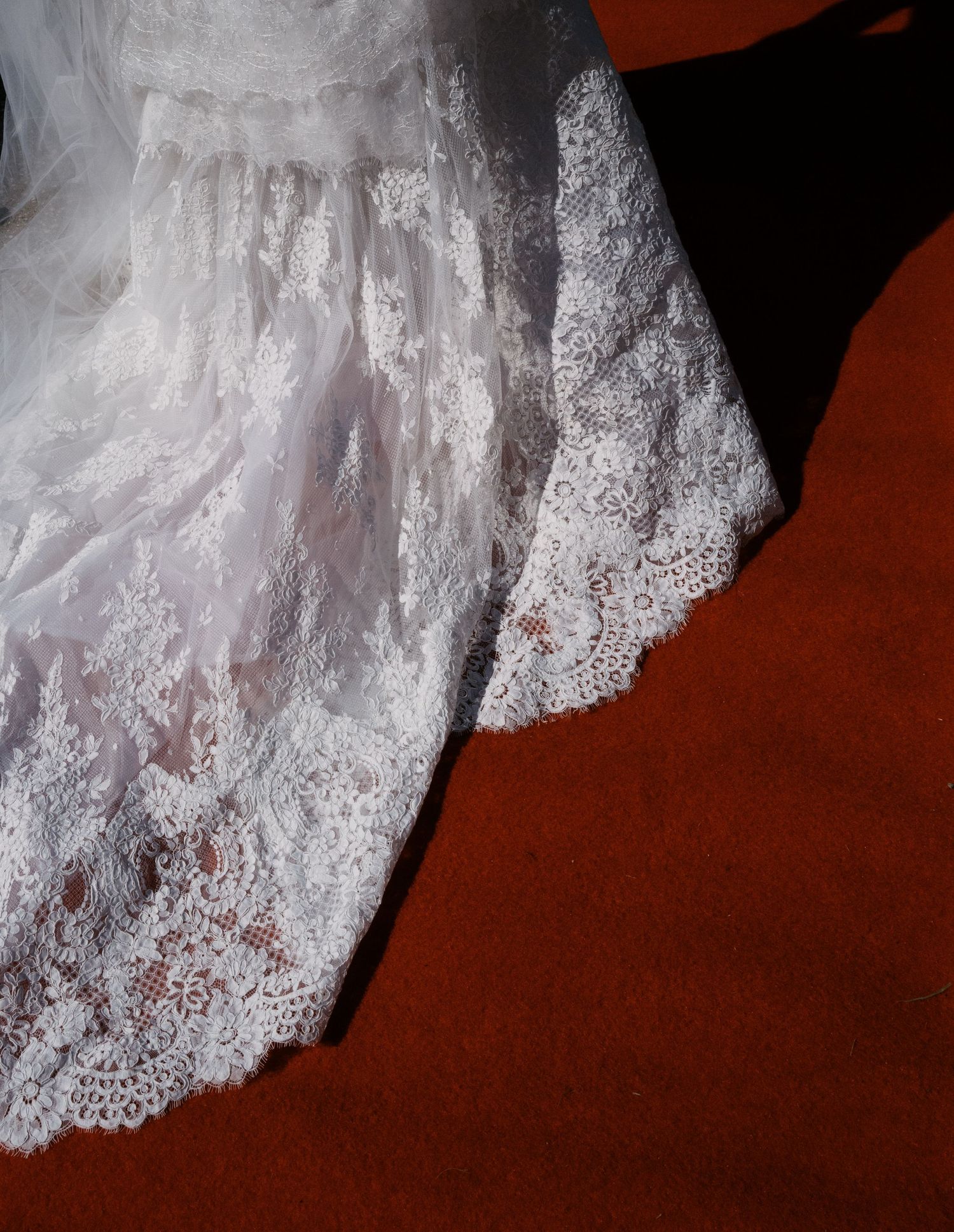 Close-up details of intricate white lace wedding dress hem against dark red flooring.