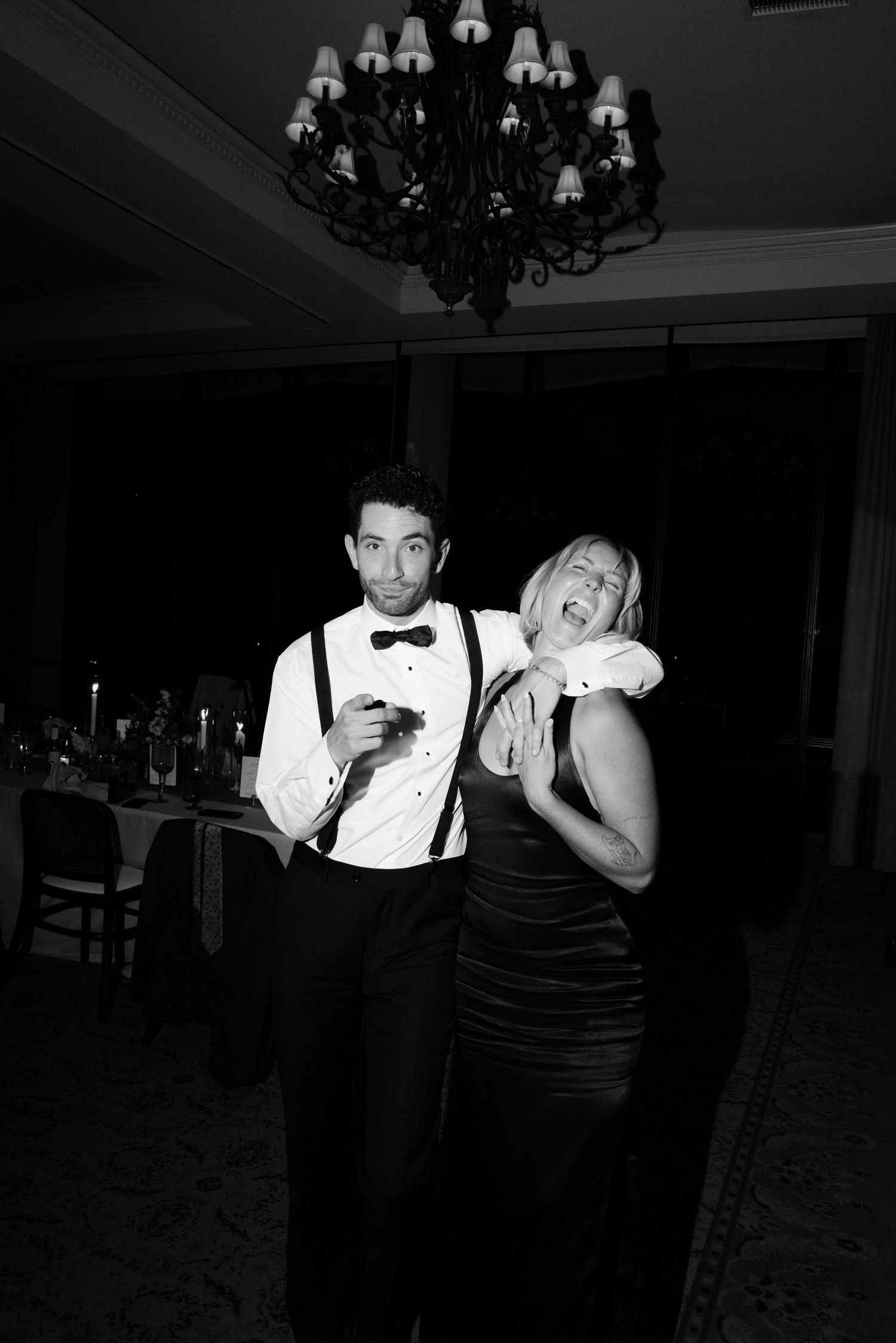 A black and white photo of a formal party with two people in elegant attire laughing under a chandelier.