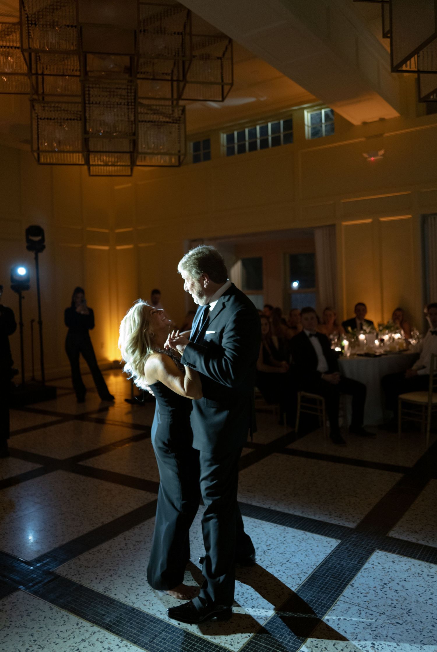 A couple performs an elegant tango dance sequence in a dimly lit ballroom with geometric lighting fixtures overhead.