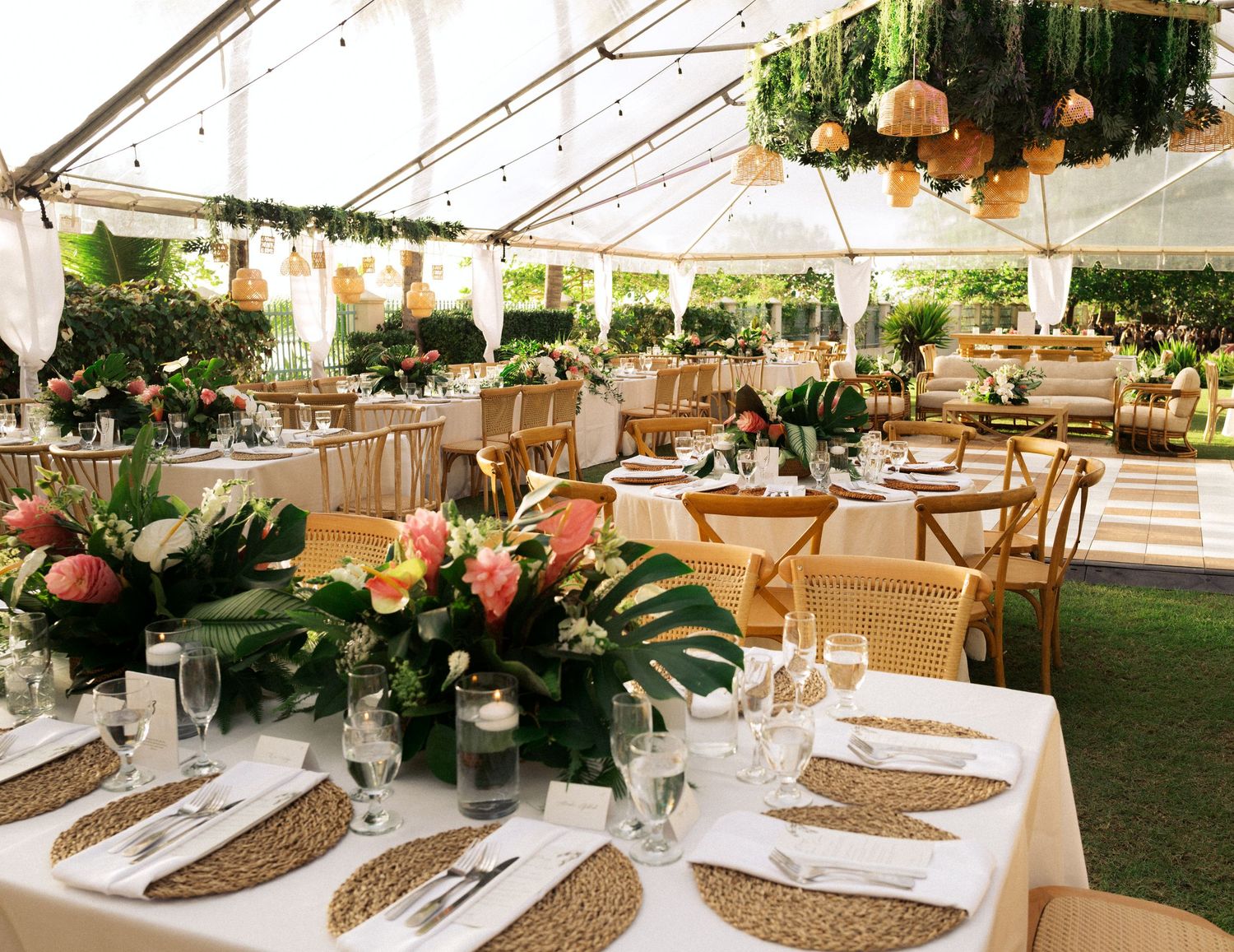 Elegant outdoor wedding reception tables decorated with tropical greenery and burlap runners under a white tent canopy.