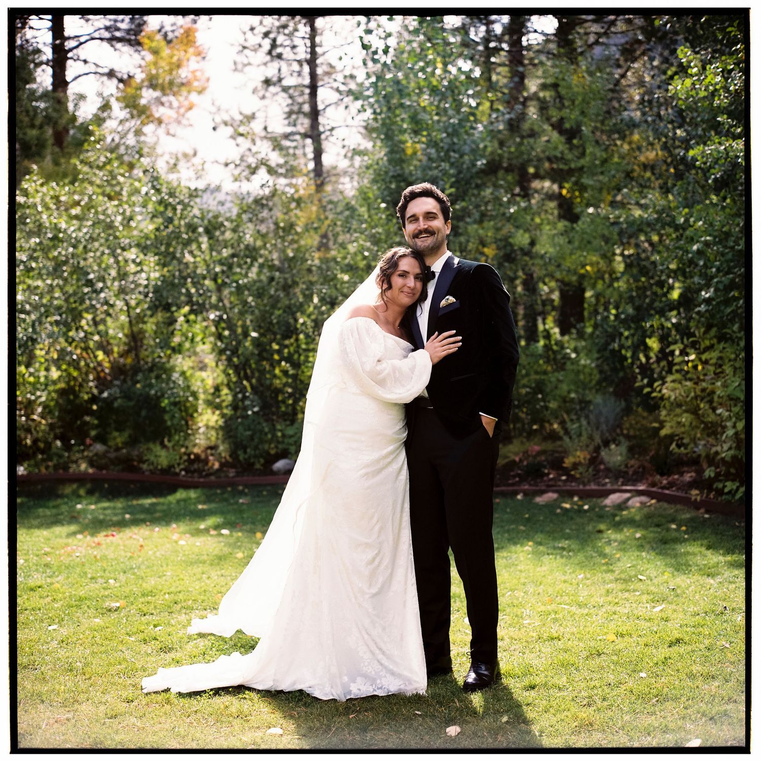 A couple in wedding attire embraces outdoors in a natural setting with trees and greenery in the background.