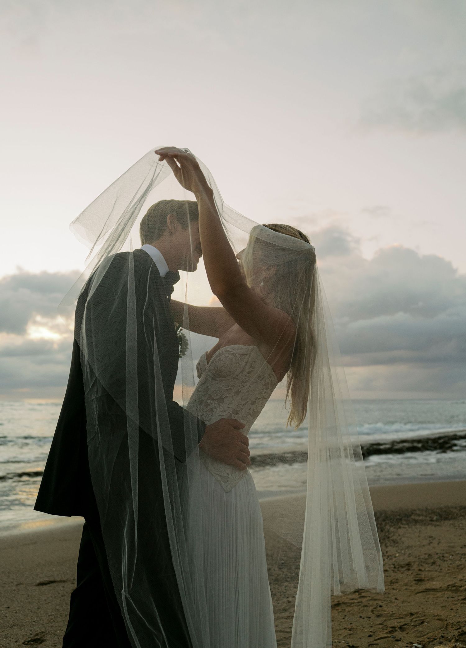 A romantic beach wedding photo silhouetted against a cloudy sunset with bride and groom sharing an intimate moment.