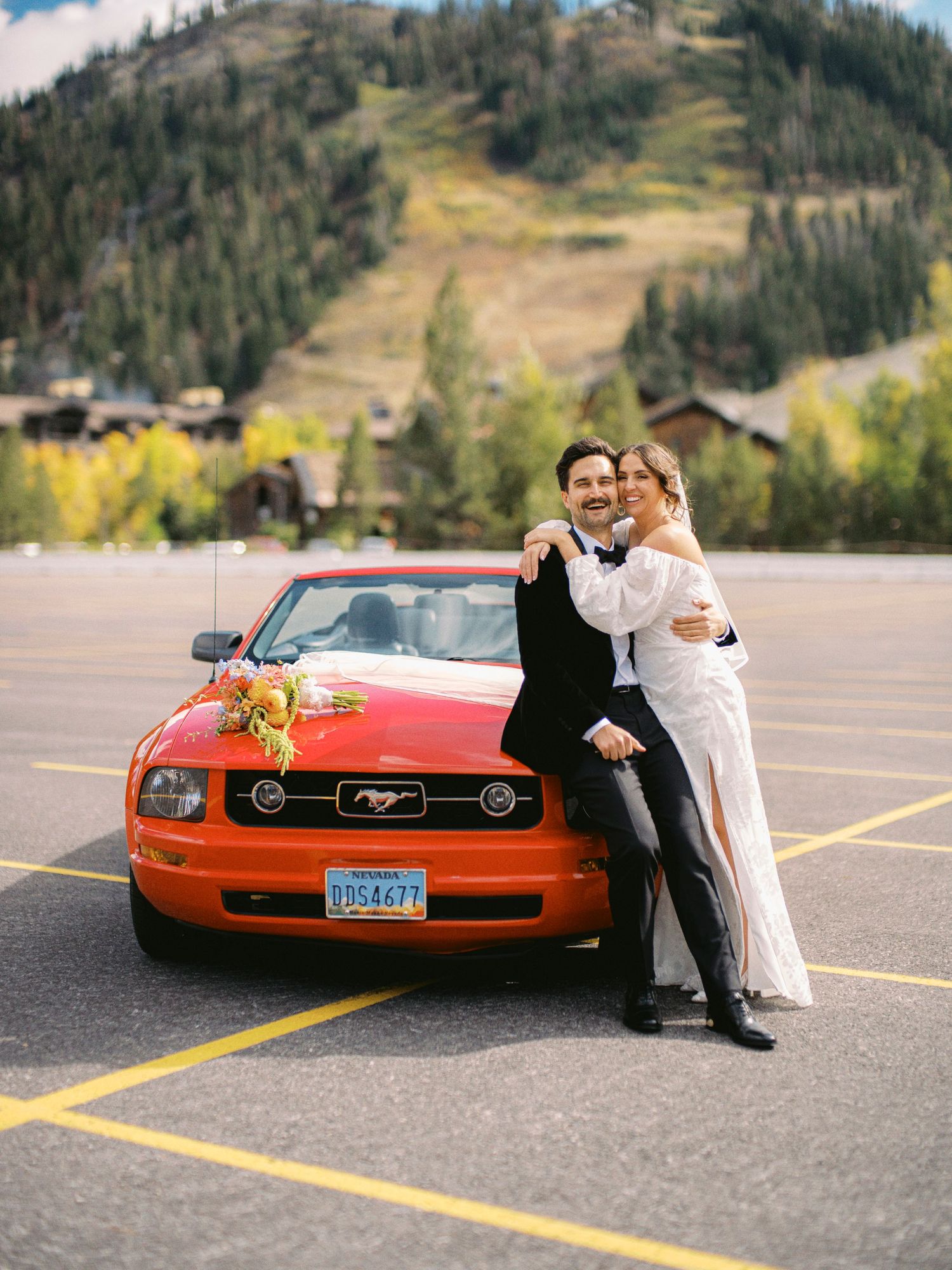 A couple poses with a bright red classic car in a mountain parking lot.