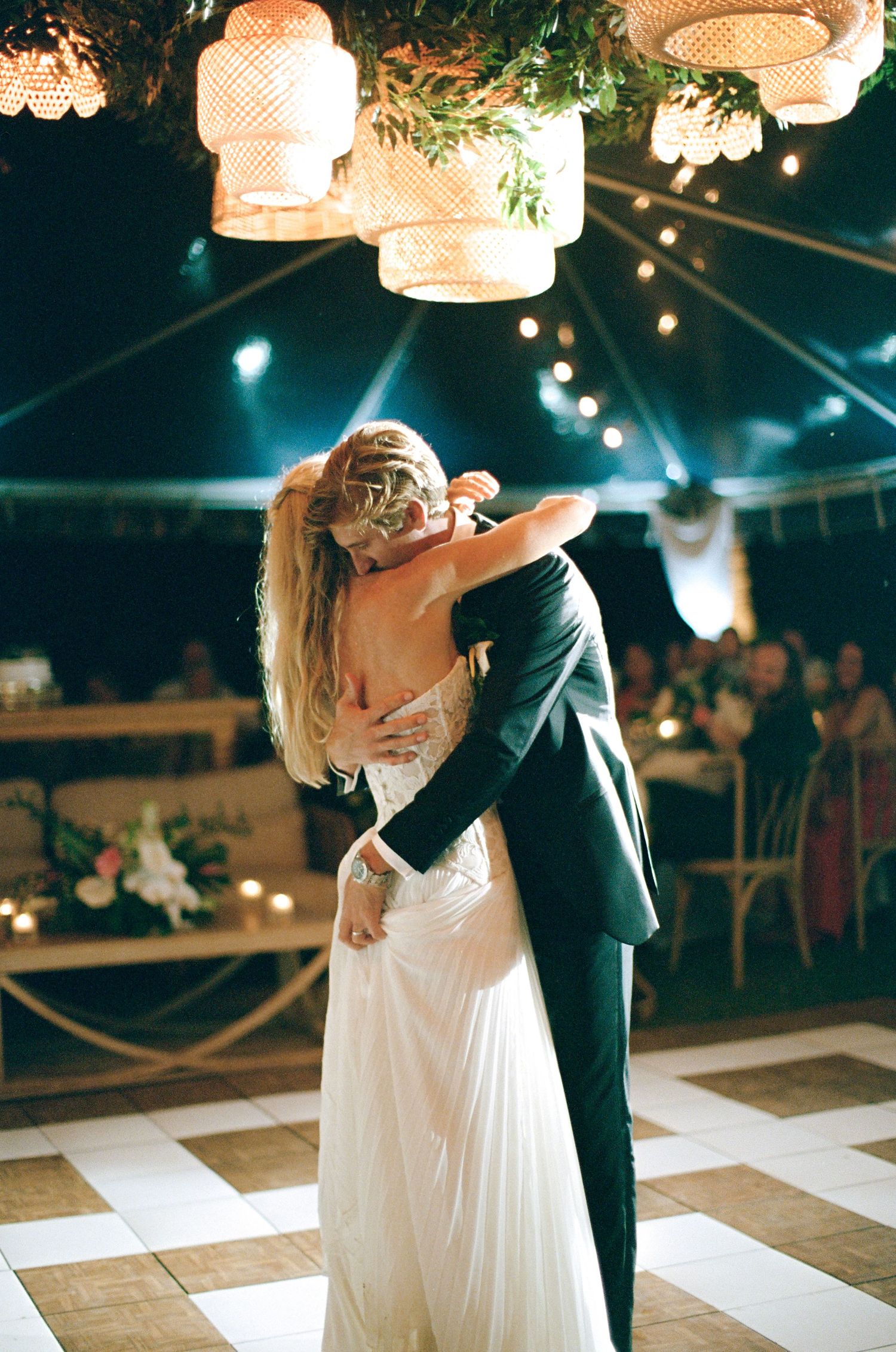 A couple shares their first dance at a wedding reception under woven basket pendant lights on a checkered dance floor.