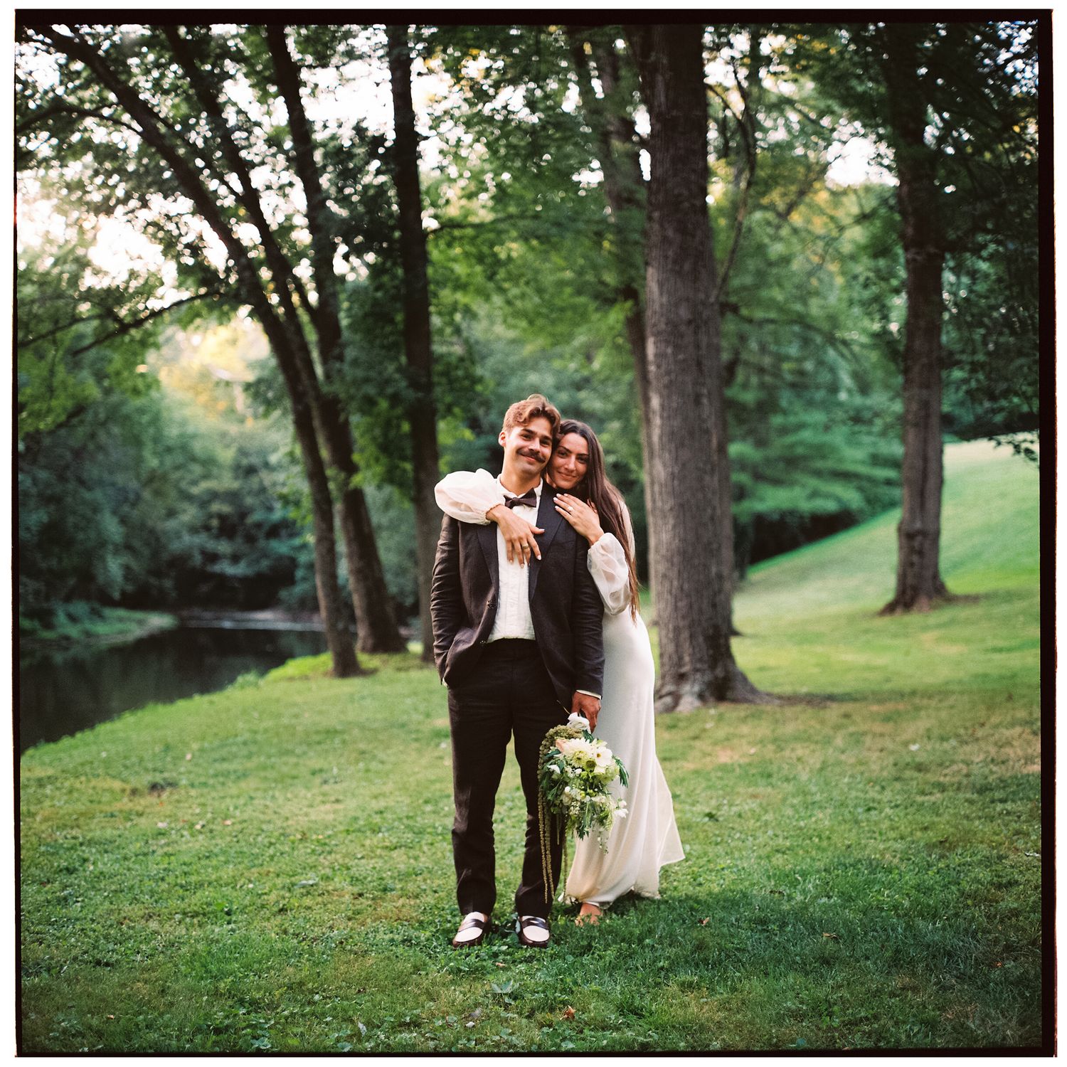 A romantic outdoor wedding scene unfolds beneath tall trees on a grassy hillside.