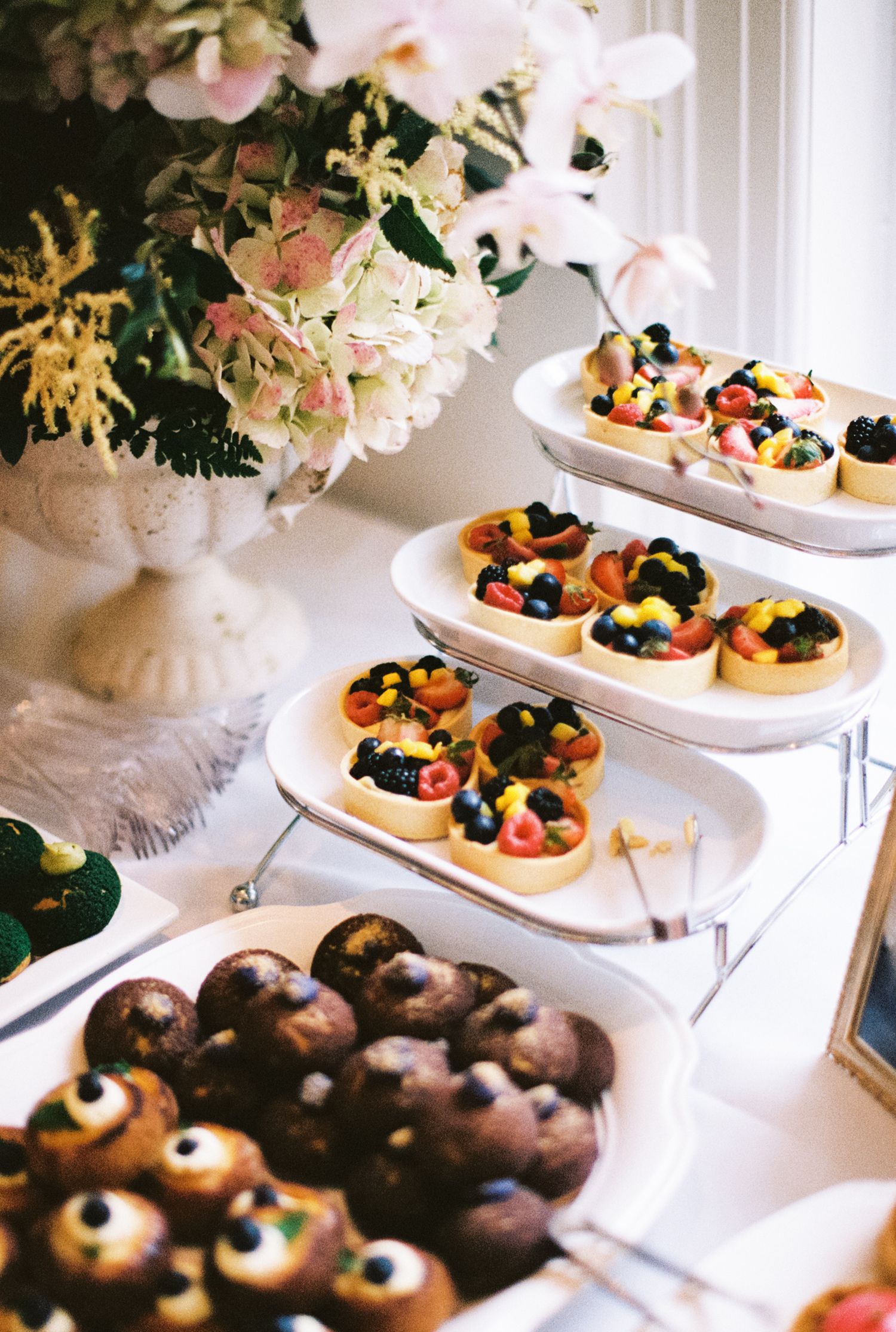 Elegant dessert table display featuring colorful fruit tarts and chocolate donuts arranged with floral decorations.