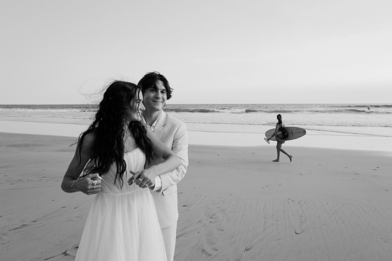 A romantic beach wedding scene with people walking along the shoreline in formal attire.