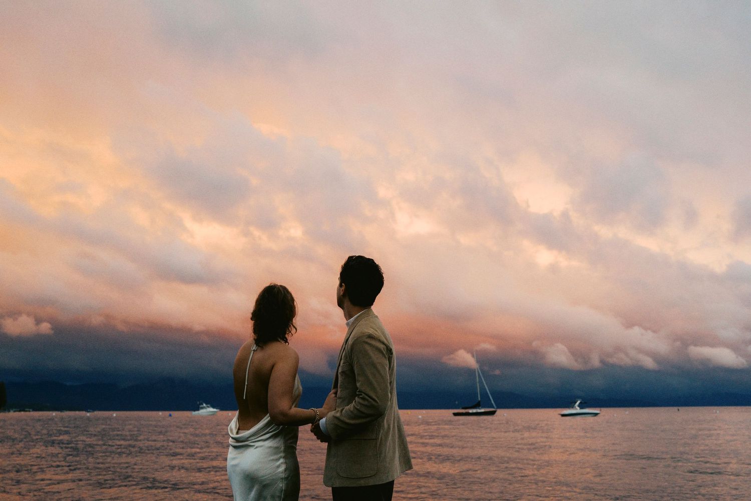 Two silhouettes stand together watching sailboats on a lake at sunset with dramatic orange and purple clouds above.