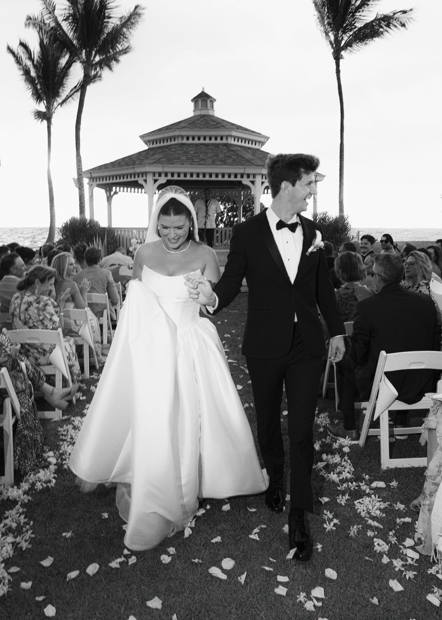 A black and white wedding photo showing a couple exiting their ceremony with a gazebo and palm trees in the background.