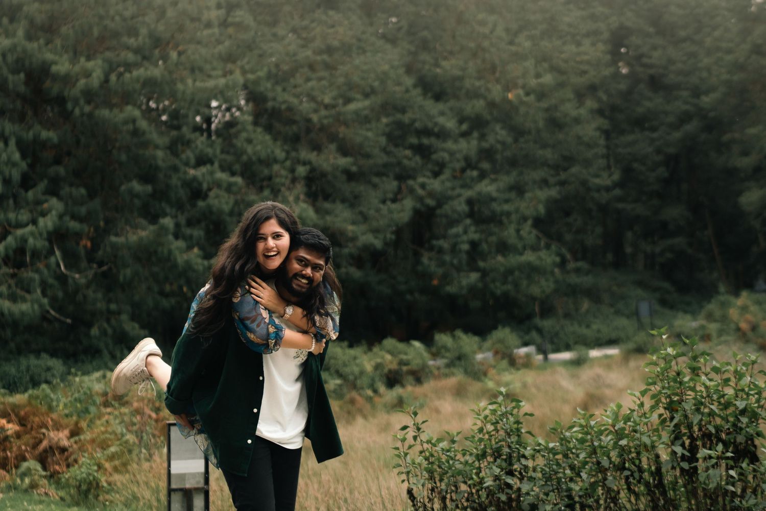 A playful couple embraces in an outdoor field with forest in background.