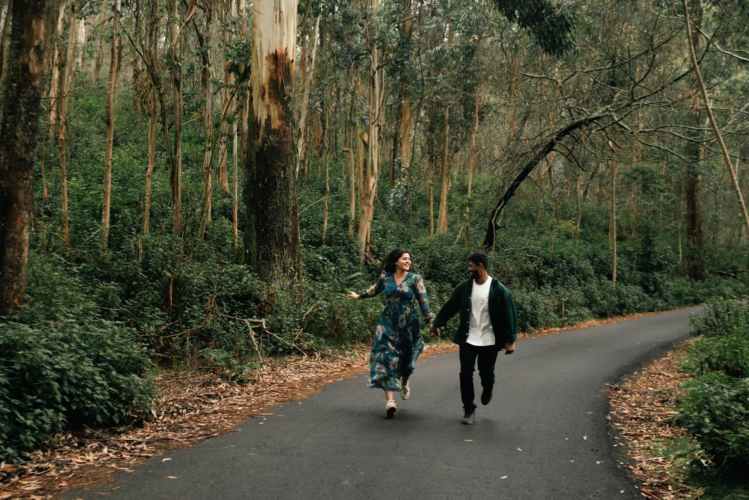 A couple running together on a forest path surrounded by tall trees.