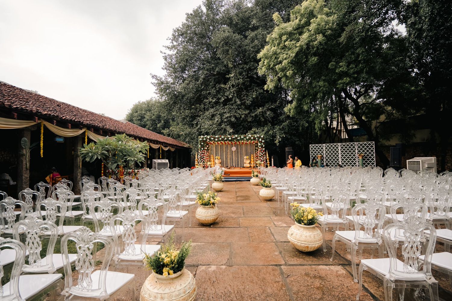 Outdoor wedding venue setup with white chairs and decor under tall trees.