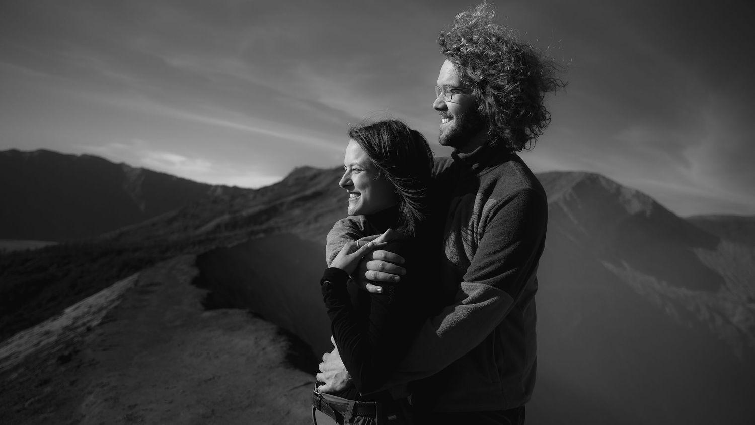 Two figures stand embracing on a mountain ridge against a dramatic black and white sky and mountain landscape.