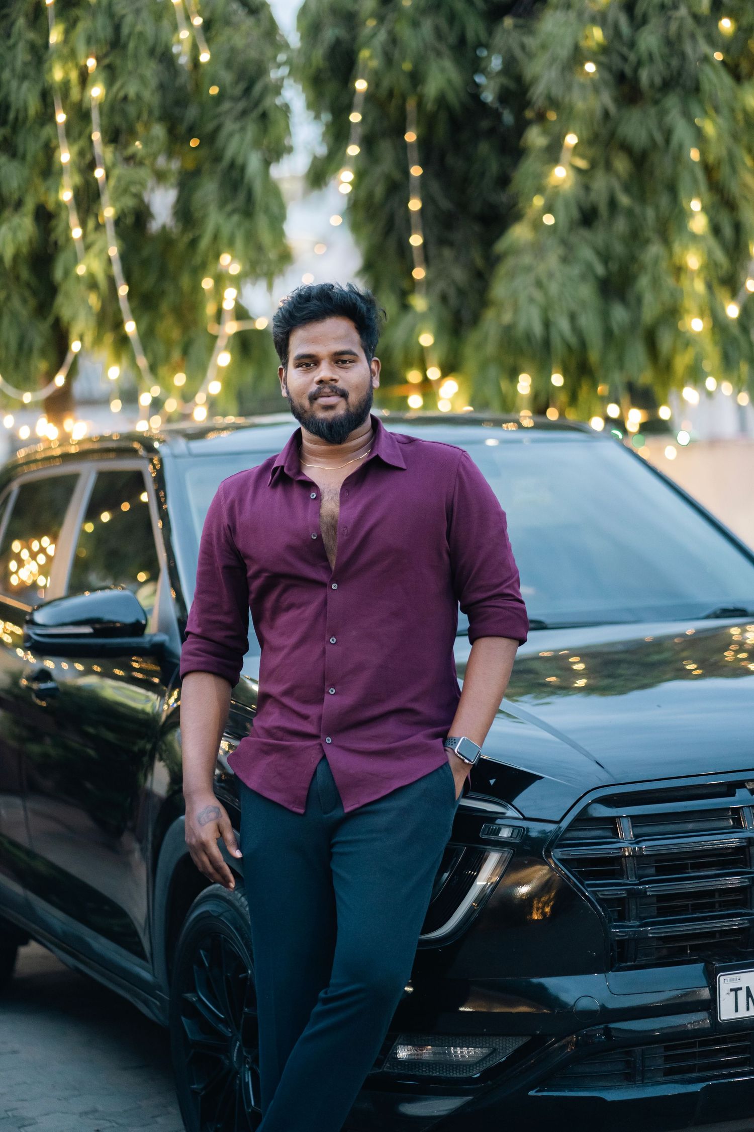 A person in a burgundy shirt and dark pants stands in front of a black car with decorative lights in the background.