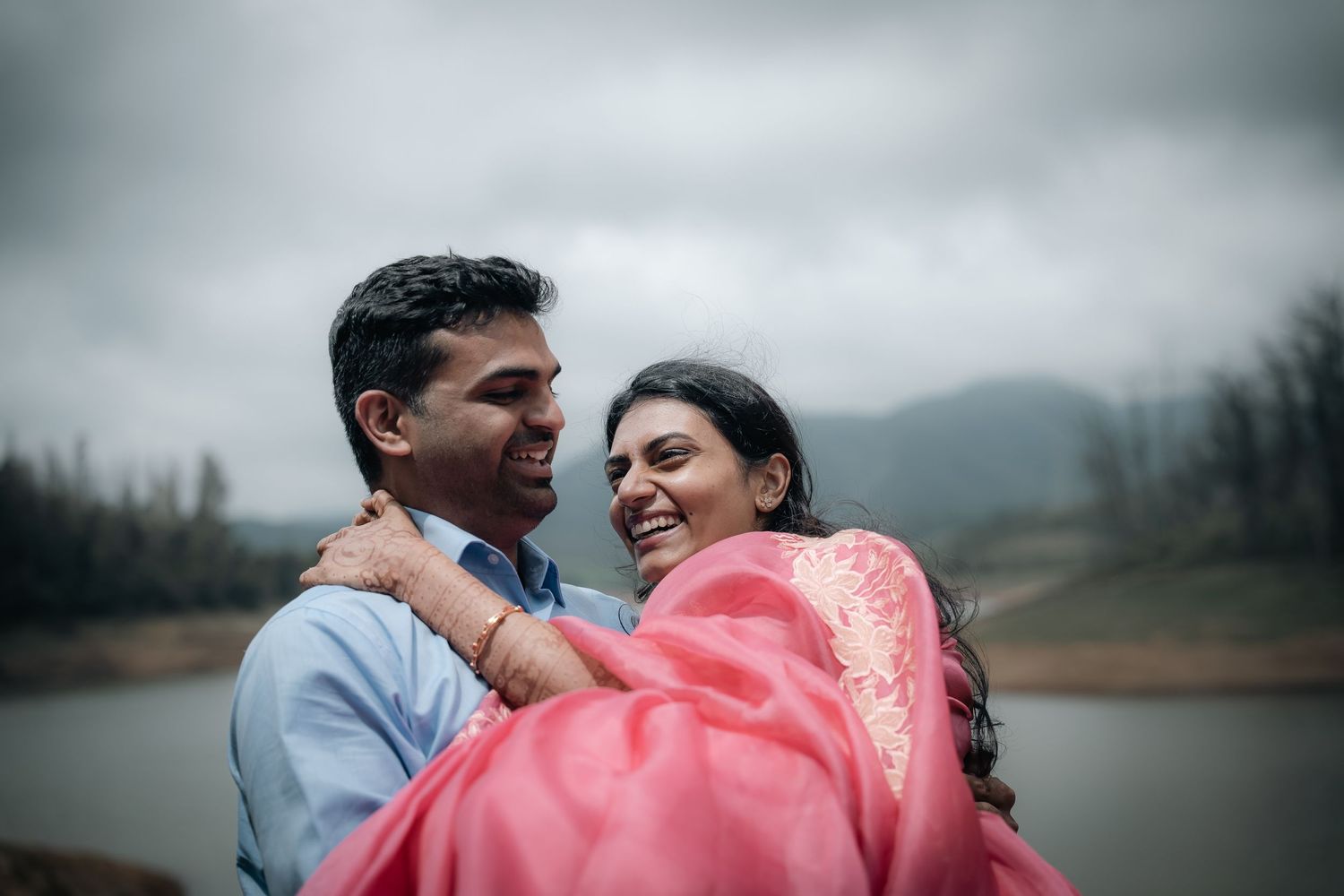 Couple embracing near waterfront with cloudy skies and mountains.