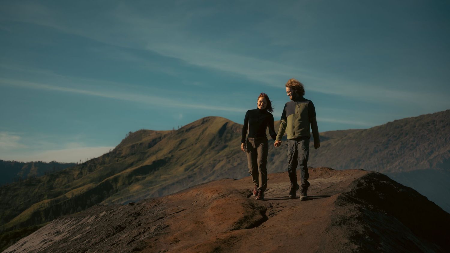 Hikers trek along a rugged mountain ridge trail against a blue sky at sunset.