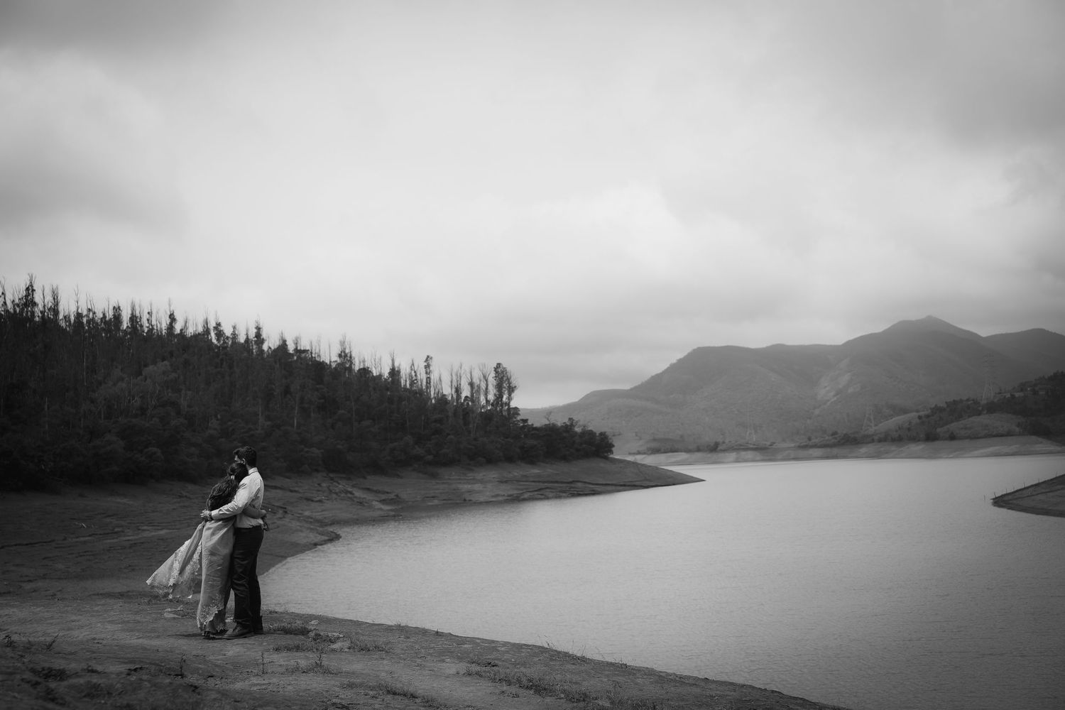 People stand by a misty mountain lake with evergreen forest in background.