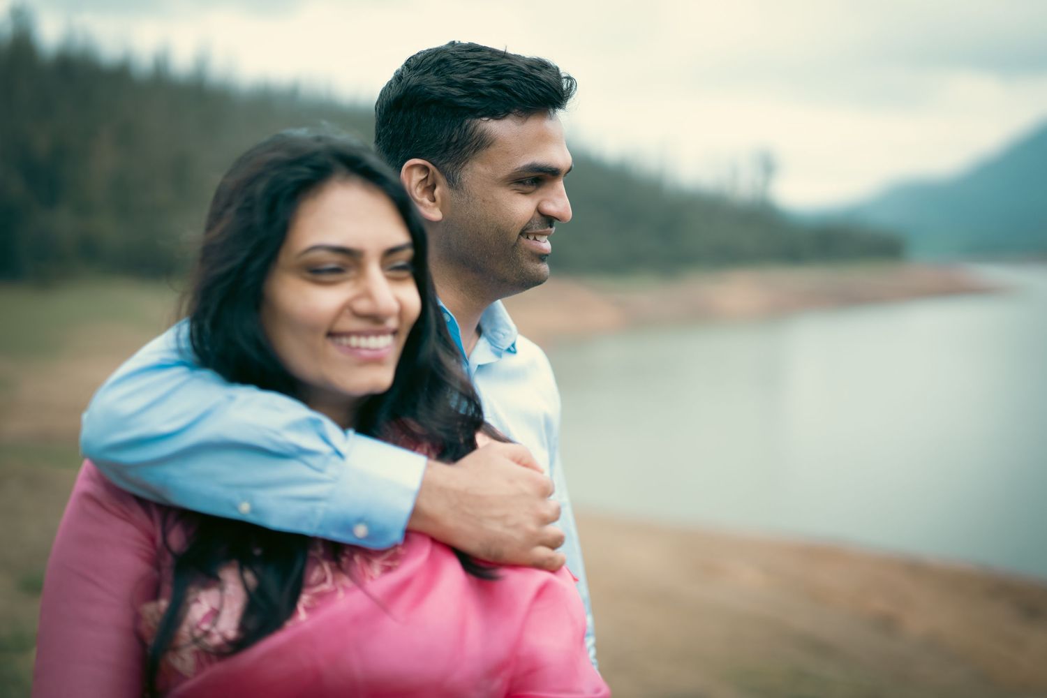 Couple embracing by a serene lake with mountainous landscape in background.