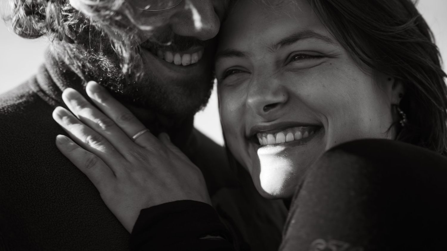 A black and white portrait of a couple laughing and embracing in natural light.