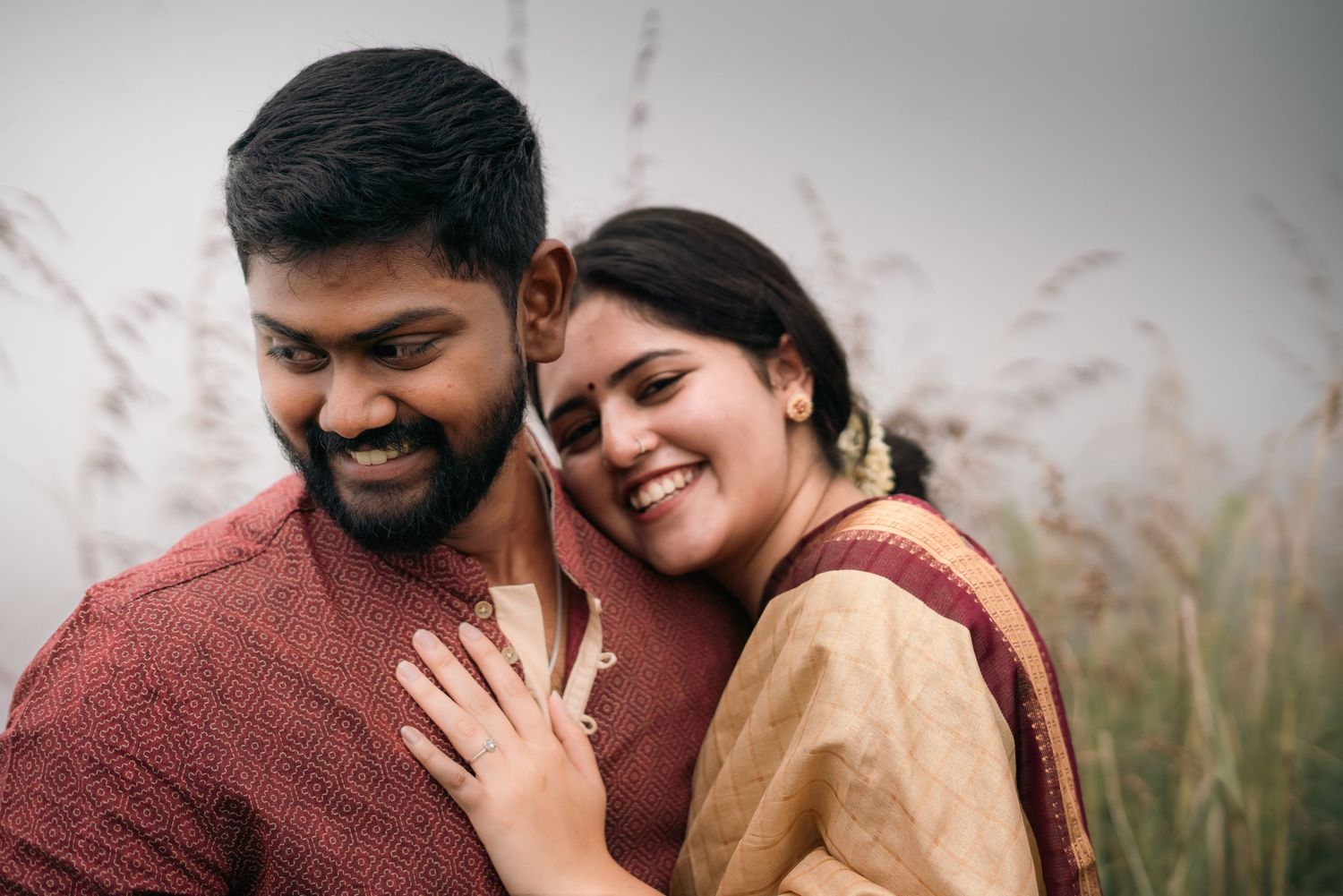 A couple smiles and embraces while wearing traditional South Asian attire against a natural backdrop.