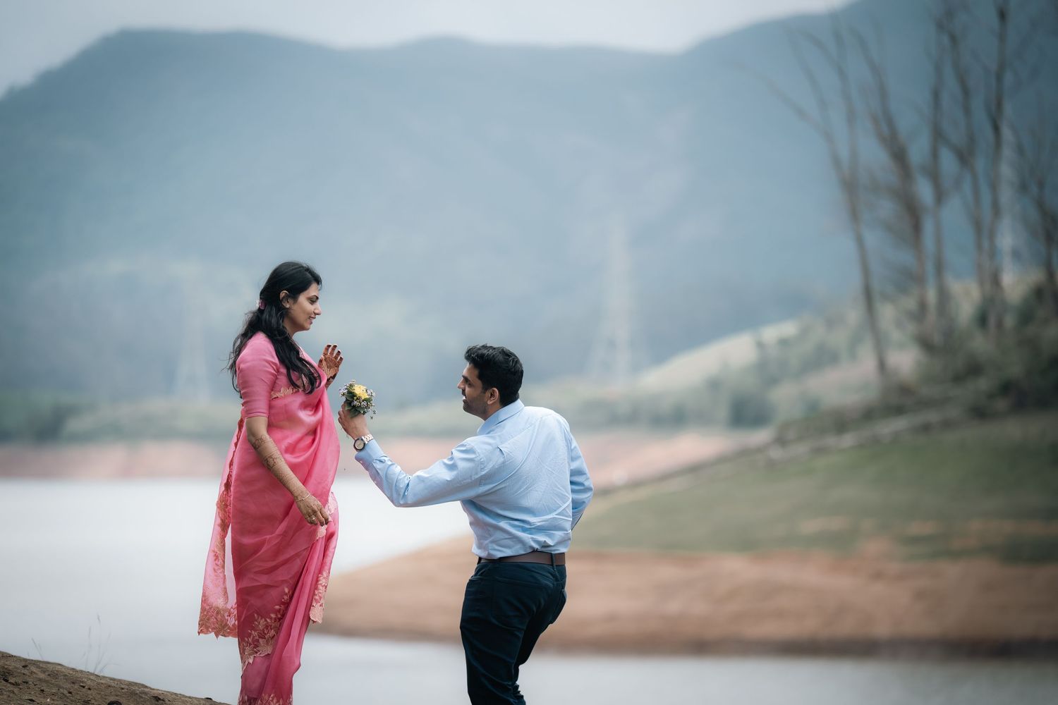 A couple in traditional Indian attire walking hand in hand near a misty lake with mountains in the background.