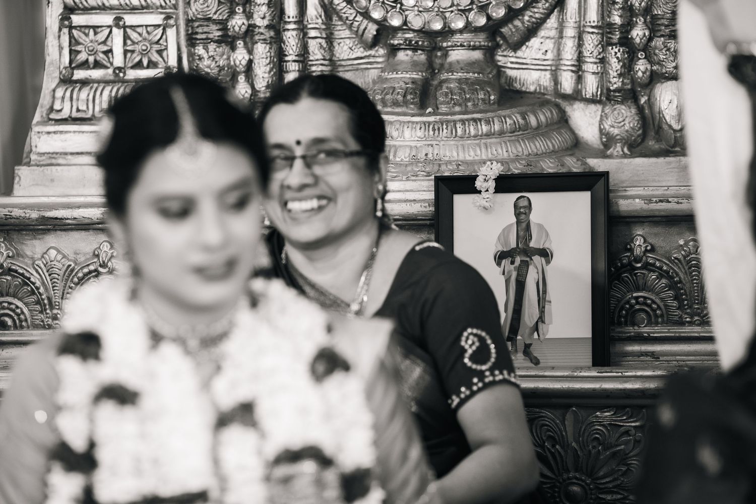 A black and white photograph shows a candid moment with two women at an elaborately decorated traditional event setting.