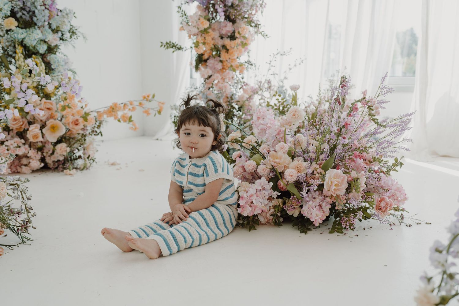 Small child in striped outfit sitting on white floor with elegant floral arrangements.