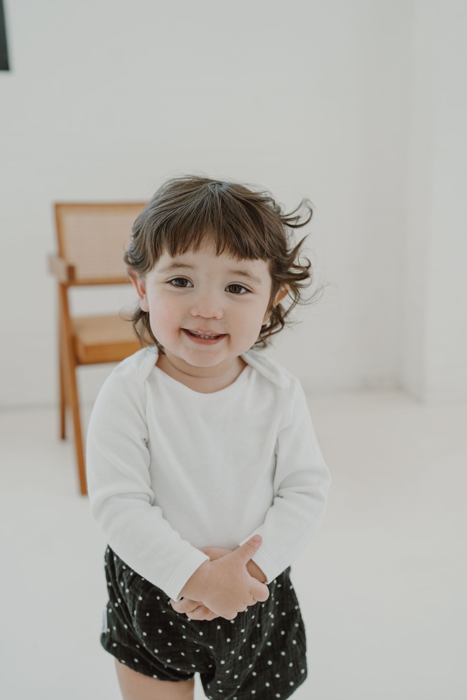 Toddler in white top and polka dot shorts smiling at camera in bright studio setting.
