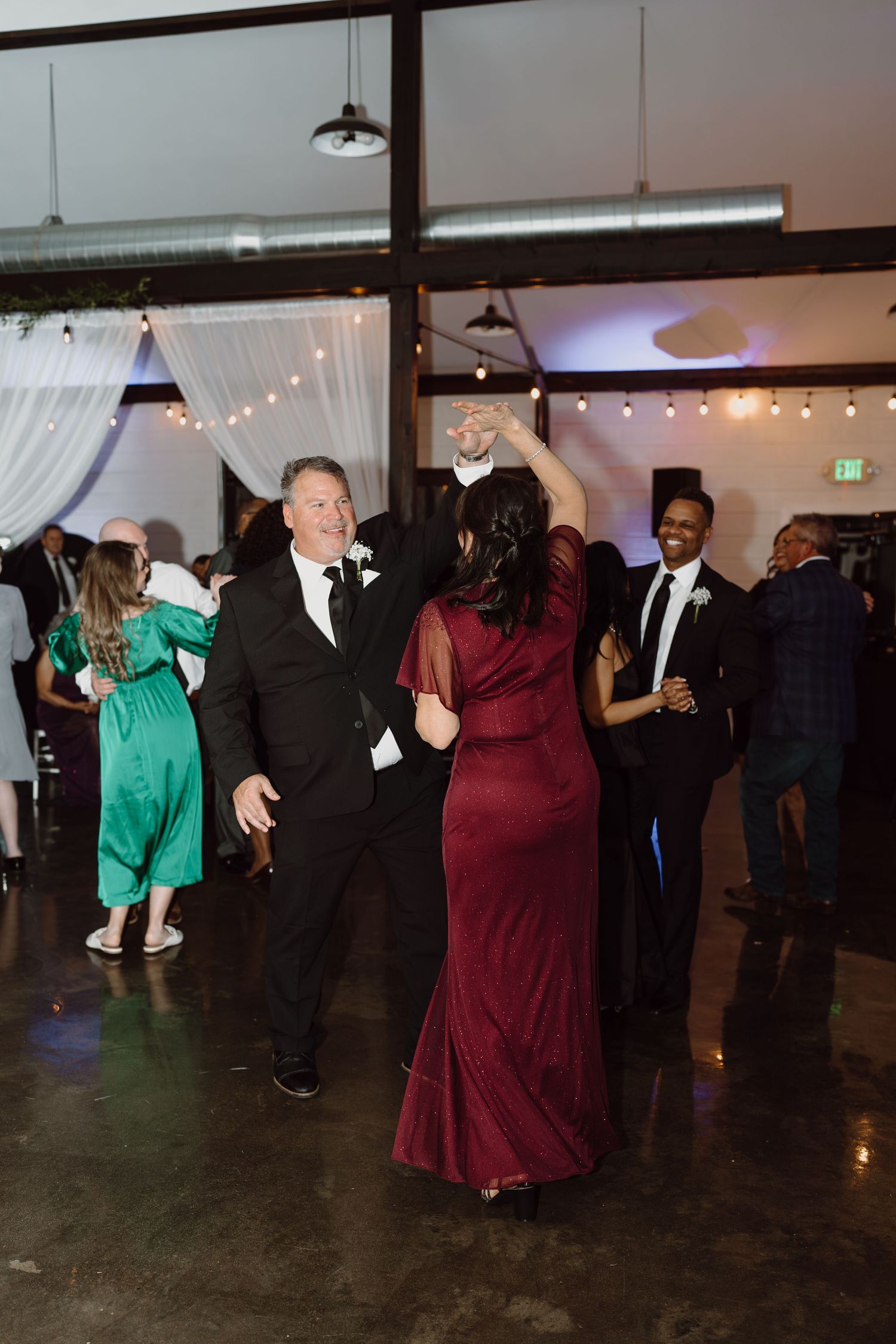 Wedding guests dancing in formal attire at reception with burgundy dress and black suits.